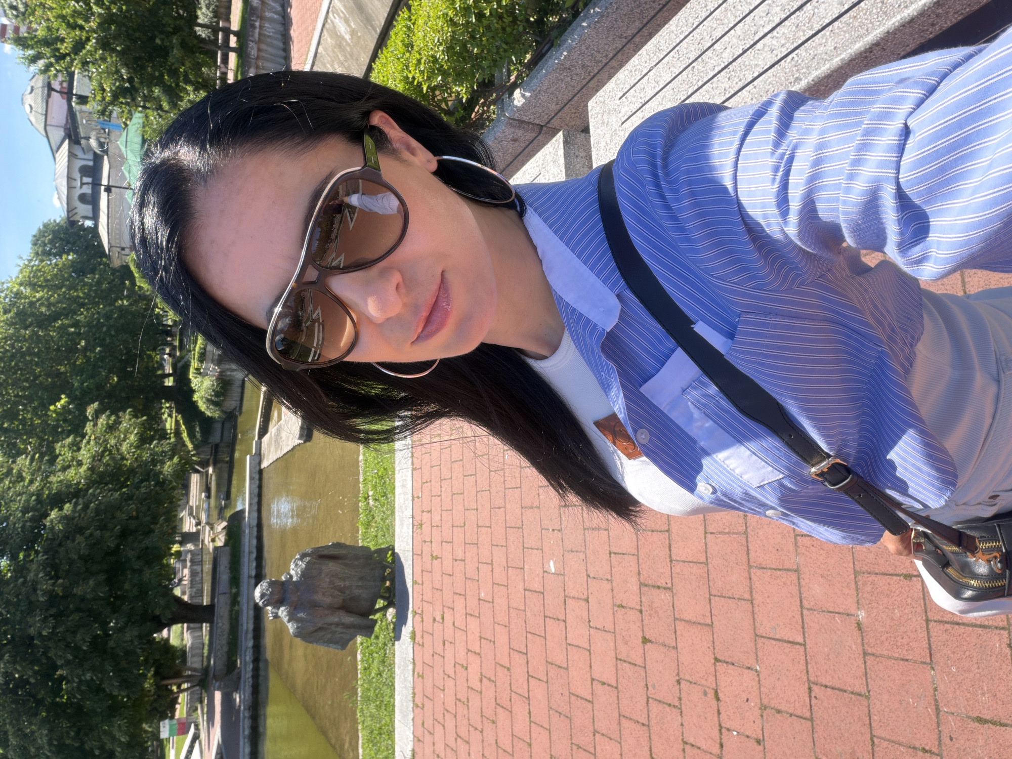 A woman with dark hair and sunglasses is taking a selfie outdoors. She is wearing a blue striped shirt, a white undershirt, and large hoop earrings. A black purse is slung across her body.
In the background, there is a body of water and a bronze statue on the shore. Red brick pavers lead away from the water. There are trees and greenery in the background. The dome of a building is visible through the trees.
The location is likely the Kaylaka Park in Pleven, Bulgaria, based on the presence of the water feature, the statue, and the architecture in the background.