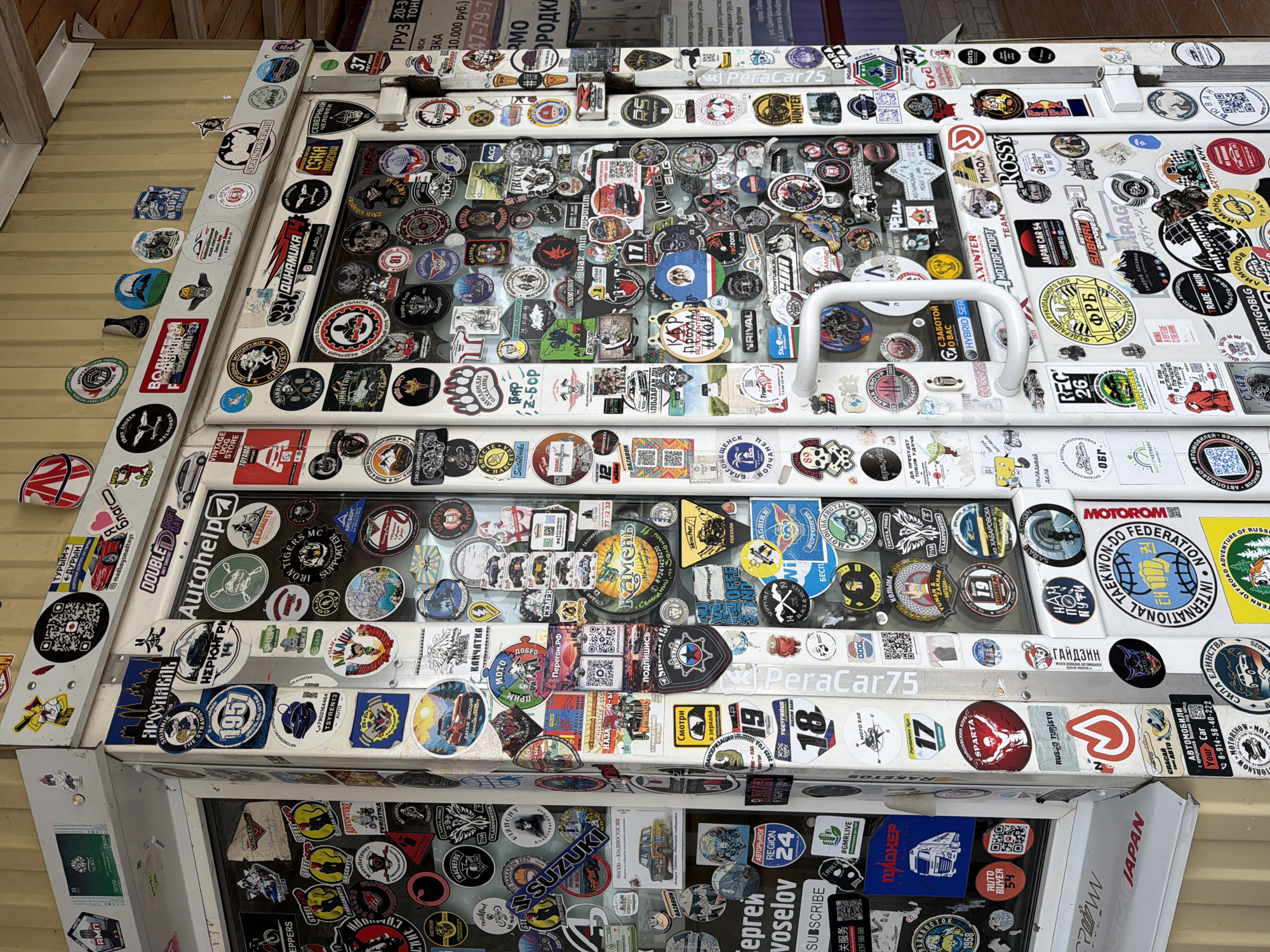 The image presents a close-up, high-angle shot of what appears to be the lid of a storage container, or a similar box-like structure, heavily adorned with a multitude of stickers. These stickers vary in size, shape, and color, and seem to represent a wide range of interests including motorsports, travel, automotive brands, local clubs, and personal affiliations. Many of the stickers contain text in Russian and other languages.
The lid is white, and is constructed of a combination of metal and possibly plexiglass. Some stickers are applied on the glass. A white handle is affixed to the lid.
In the lower portion of the image, there is an apparent continuation of the sticker-covered surfaces suggesting that the side of the container is also decorated.
The surface underneath the container lid appears to be corrugated metal paneling.
Given the mention of Кирга, Russia in the prompt, it could be inferred that the photograph was taken in that location. However, this cannot be definitively proven from the image alone.