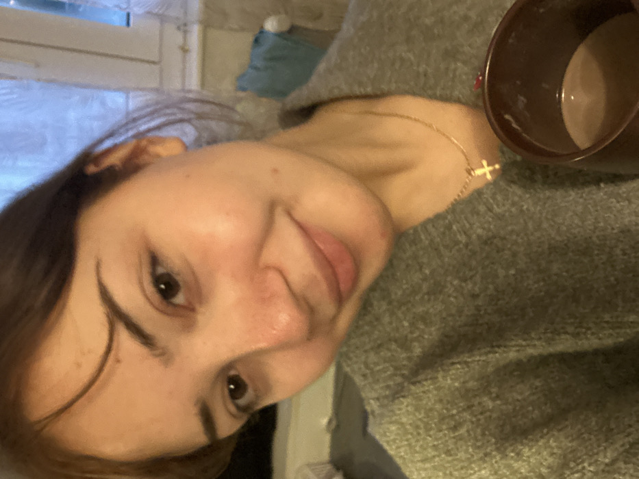 A woman poses close to the camera, with her head tilted. She has brown hair and brown eyes. She wears a gray sweater and a delicate gold necklace with a cross pendant. A brown mug, seemingly filled with coffee or chocolate, is visible in the upper right corner. A window with white blinds is in the background.
