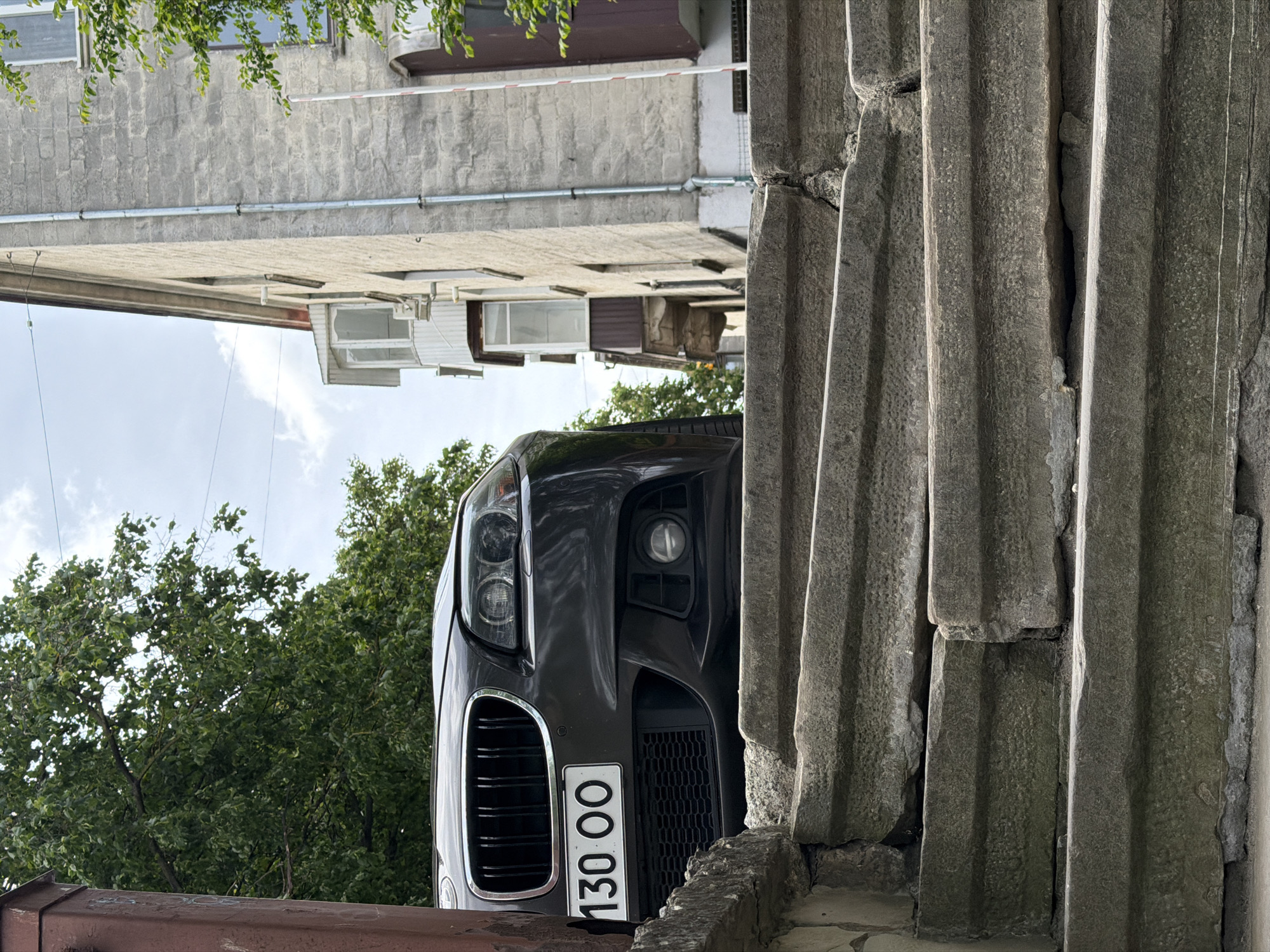 The image shows a black car with a license plate reading "130 00" partially visible behind a gray, textured concrete wall. The car appears to be parked on a street with trees in the background. In the distance, there are buildings with light-colored walls and brown window frames. The photo is taken from a low angle, looking up at the car and the buildings. The sky is visible with some clouds. The image is rotated by 90 degrees clockwise. The location is Ternopil, Ukraine.