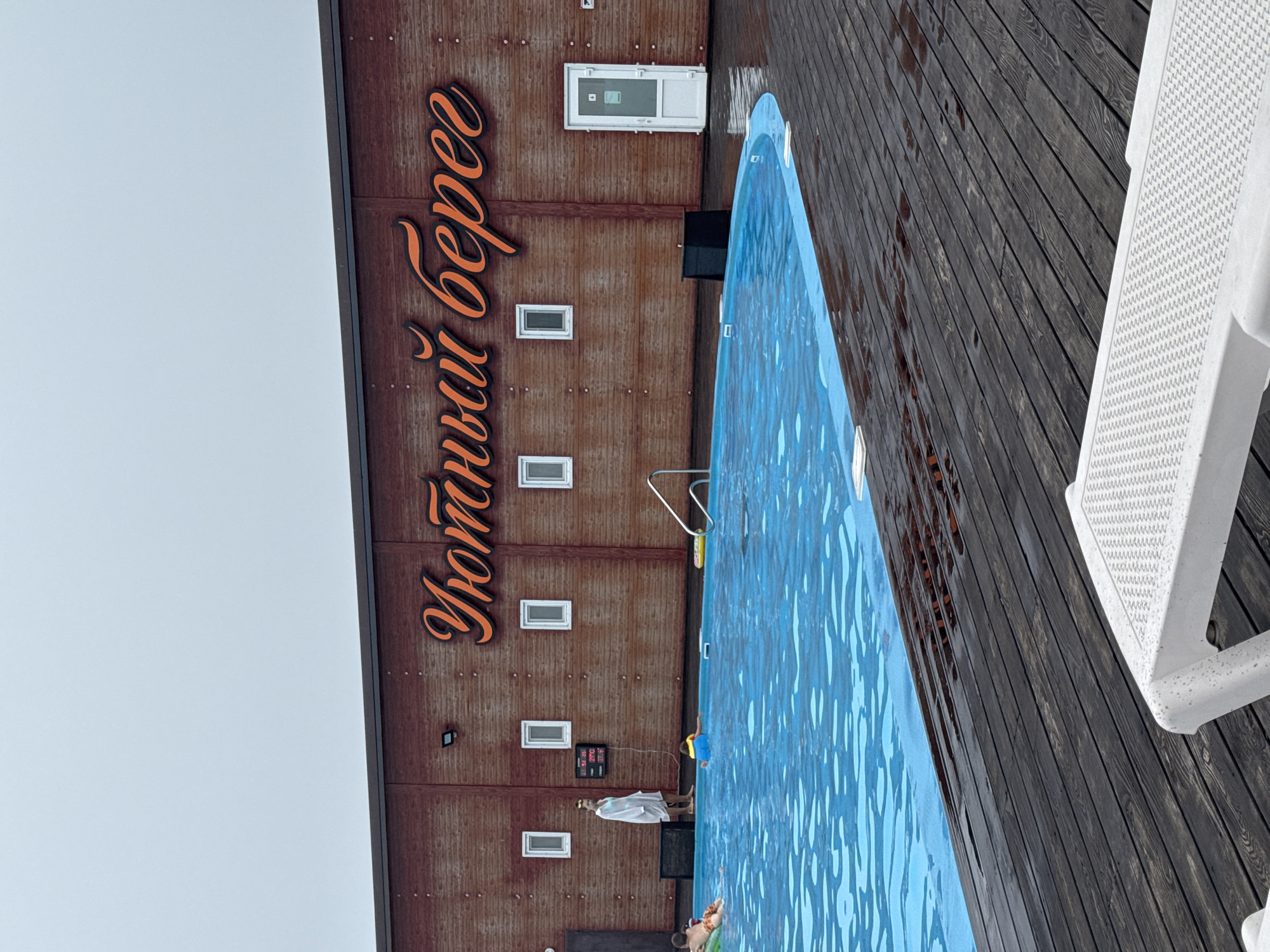An outdoor swimming pool area at a facility with wooden siding is depicted. The facility has large orange lettering that reads "Уютный берег" (Cozy Shore). A blue swimming pool is in the foreground, with a dark wooden deck surrounding it. Several people are in or near the pool. A woman in a white robe stands on the deck near a digital display. Two children are in the pool, one wearing a yellow floatation device and another on an inflatable ring. In the foreground, a white plastic lounge chair is visible, with water droplets on its surface. The sky is overcast and grey. The location is identified as Rakushka, Russia.