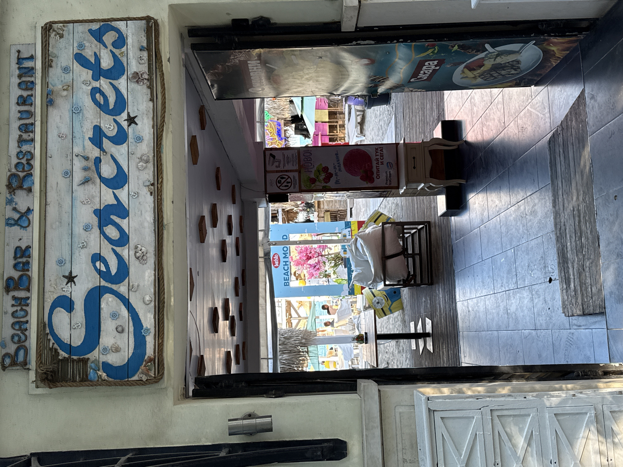 The image shows the entrance to a beach bar and restaurant called "Secrets." A weathered wooden sign with blue lettering and nautical decorations hangs above the entrance. Through the open doorway, a sandy beach area with tables, chairs, and umbrellas is visible. There are a few people scattered around the beach, some seated at tables and others walking. To the right of the entrance, a tall sign displays images of ice cream and what appears to be food. The floor inside the entrance is tiled, and the ceiling has a grid of wooden panels. The overall atmosphere suggests a relaxed, beachfront establishment.