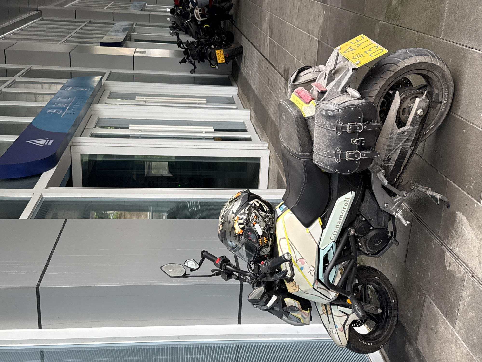The image shows two motorcycles parked on a tiled surface next to a modern building with glass windows. The primary motorcycle, positioned closer to the viewer, features a distinctive helmet placed on the handlebars and custom paintwork with the word "Cinnamoroll" displayed on its side. A leather saddlebag is attached to the rear. The license plate of this motorcycle is partially visible. Behind it, a second motorcycle is parked, with a license plate that reads "渝A V672M" and "渝A 031YE." The architecture of the building suggests a contemporary design, with repeating windows and metal framework. A blue sign with Chinese characters is fixed to the building's exterior. The scene is located in Wulong District, China.