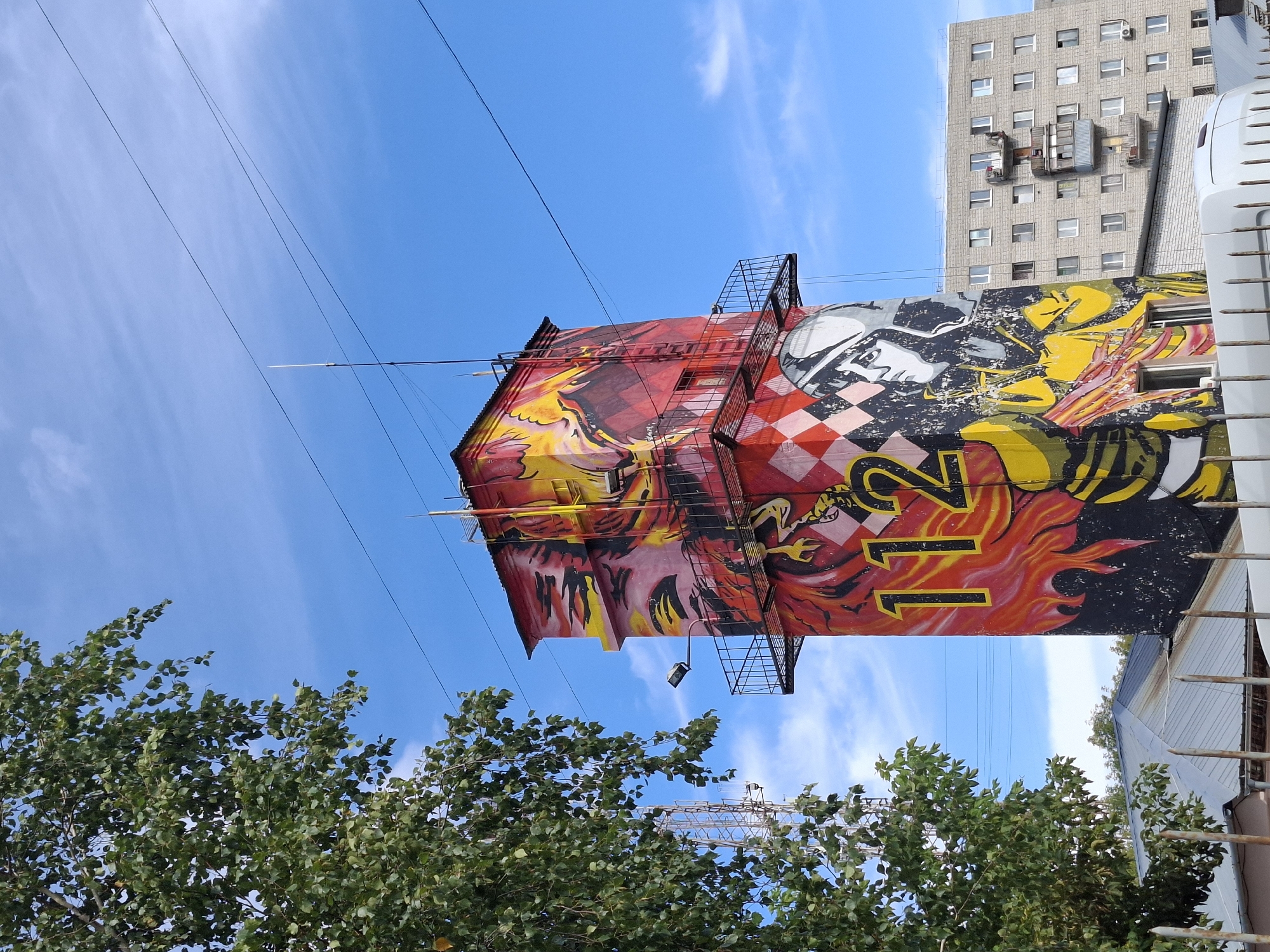 A tall, rectangular tower, adorned with vibrant graffiti, stands against a clear blue sky with scattered white clouds. The graffiti features bold imagery including flames, the number "112" in large yellow numerals, and a stylized portrait of a person wearing a helmet. The tower has a red roof and external metal stairs and platforms. To the left, a large tree with green leaves partially obscures the view. In the background, a modern, multi-story apartment building with numerous windows is visible, along with power lines crisscrossing the sky. The lower portion of the image shows the corrugated metal roof of a smaller building and a metal fence. This image was taken in the city of Tyumen, Russia.