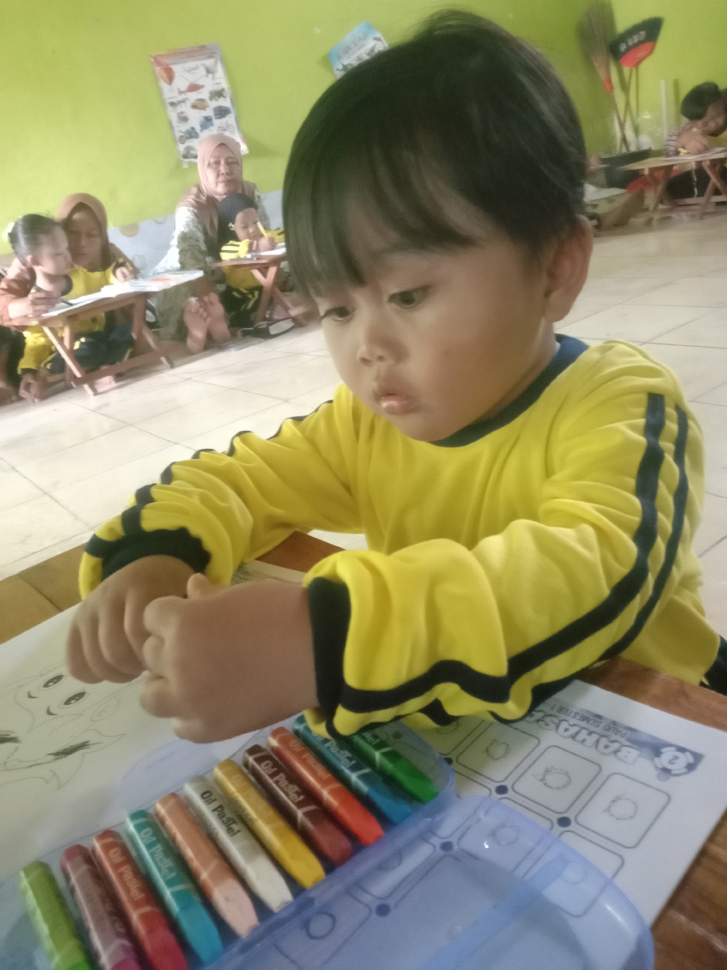 In a classroom setting, likely in Sambongrejo, Indonesia, a young child wearing a yellow long-sleeved shirt with black trim is the primary subject. The child is seated at a wooden table, focused on a drawing or coloring activity. In front of the child is an open box of oil pastels, with several colors visible, and a sheet of paper with what appears to be a partially drawn cartoon character. The child's hands are resting on the paper, indicating engagement with the activity. In the background, other children and an adult, possibly a teacher, are also present, engaged in their own activities at separate tables. Educational posters are visible on the lime green wall. The flooring appears to be tiled. The overall atmosphere suggests a learning environment for young children.