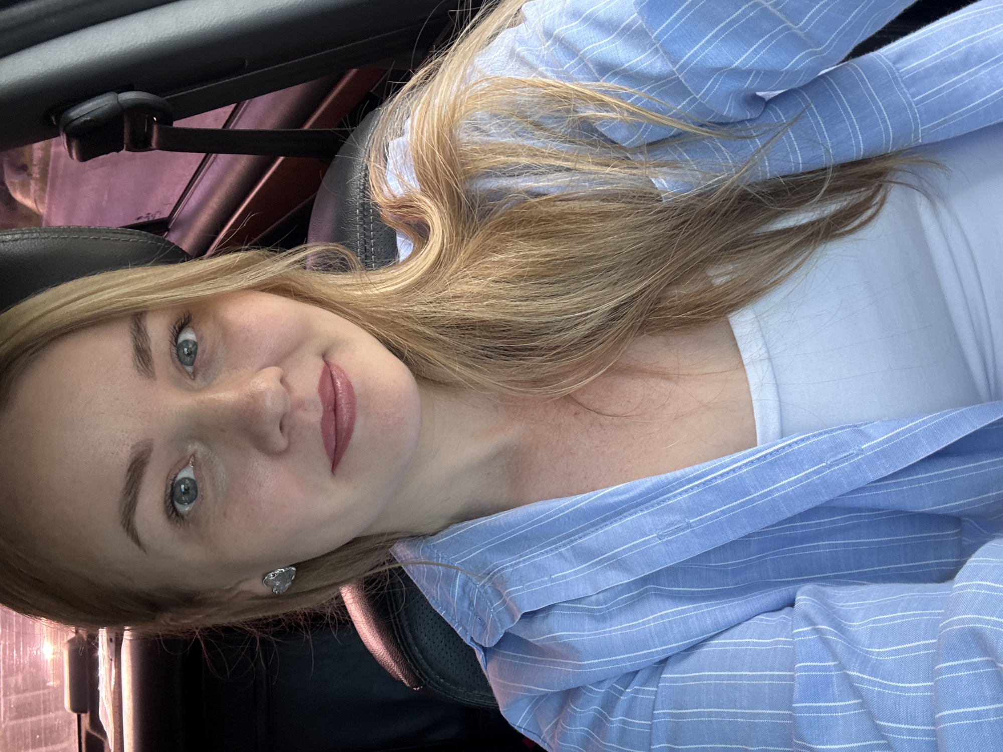 A medium shot shows a Caucasian woman with long blonde hair and blue eyes, wearing a light blue and white striped button-down shirt over a white tank top. She is seated in a car, as evidenced by the black leather seat and a seatbelt visible in the background. Her right ear is adorned with a silver, heart-shaped earring. The lighting suggests it may be daytime. No specific landmarks within Chaikovsky, Russia are visible in this image.