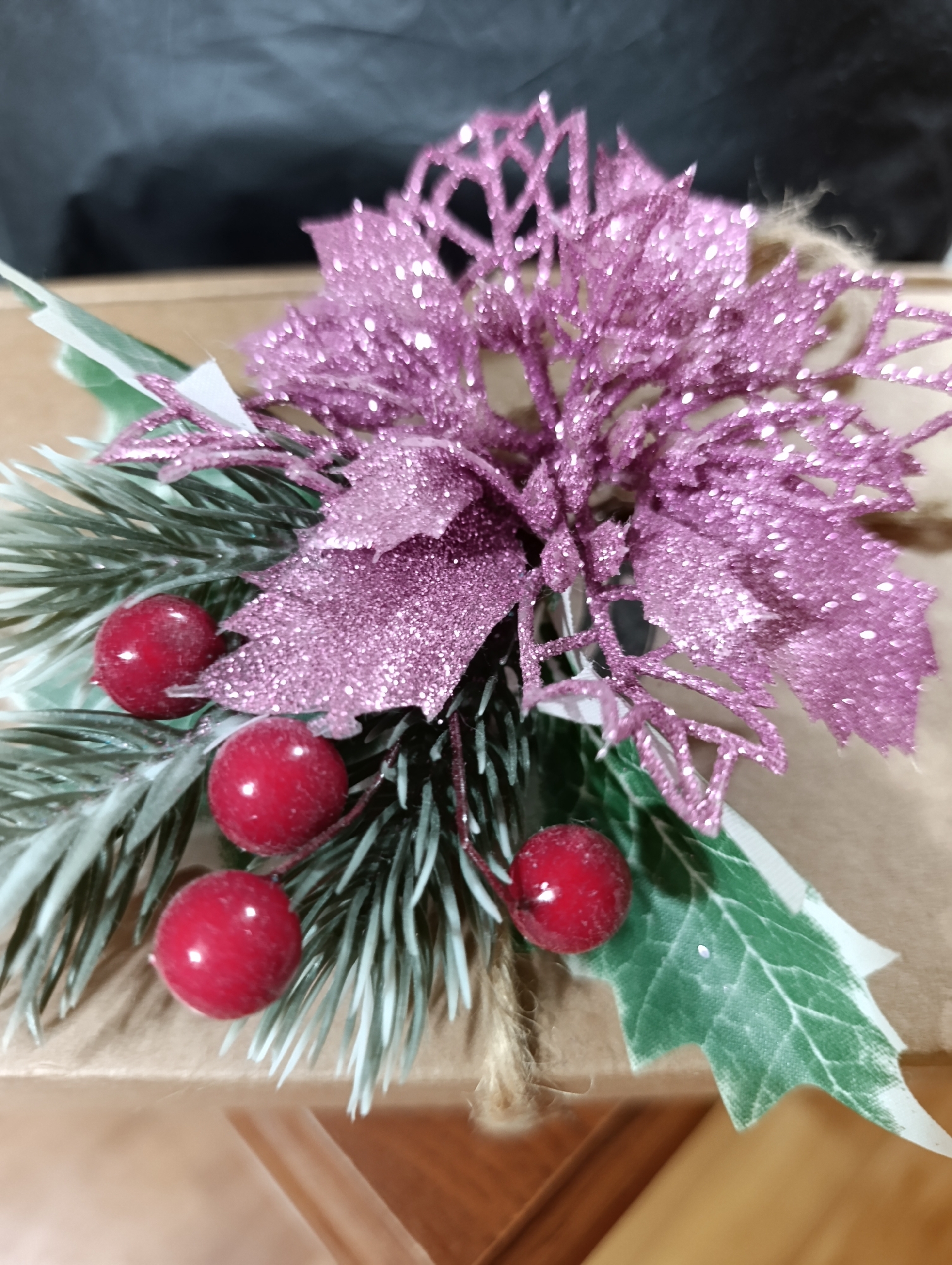 A close-up, slightly angled shot focuses on a festive Christmas decoration atop a kraft paper-wrapped gift box. The decoration consists of a glittery pink poinsettia-like flower with intricate, laser-cut petals. Nestled amongst the flower are sprigs of faux evergreen foliage, dusted with a light coating of artificial snow. Several bright red faux berries are strategically placed, adding a pop of color. A piece of twine is visible, wrapped around the base of the gift. The background is softly blurred, with the top portion appearing dark, possibly a wall or backdrop, and the lower portion showing a wooden surface, potentially a table or shelf. The scene is static, with no people or animals present. The overall impression is one of a handcrafted holiday embellishment ready for gifting. The location is not discernible from the visual information.