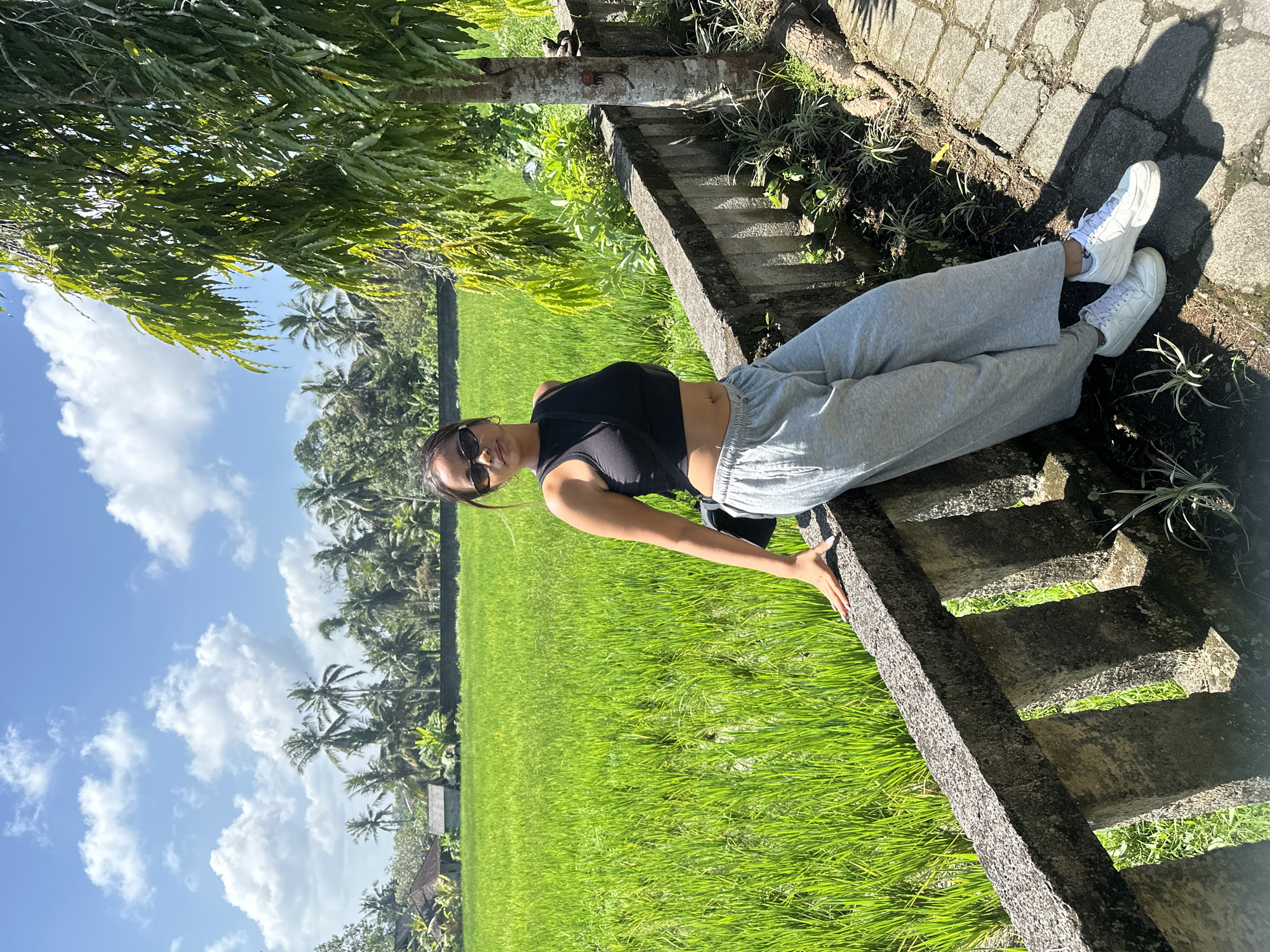 A young woman with dark hair and sunglasses is standing and leaning on a low concrete wall in front of a vibrant green rice paddy in Ubud District, Indonesia. She is wearing a black crop top, grey sweatpants, and white sneakers. Behind her, the rice field stretches towards a line of palm trees and some small buildings. The sky is a bright blue with scattered white clouds. A tree with feathery green leaves grows to the right of the woman. The ground beside the wall is paved with irregular stone blocks.