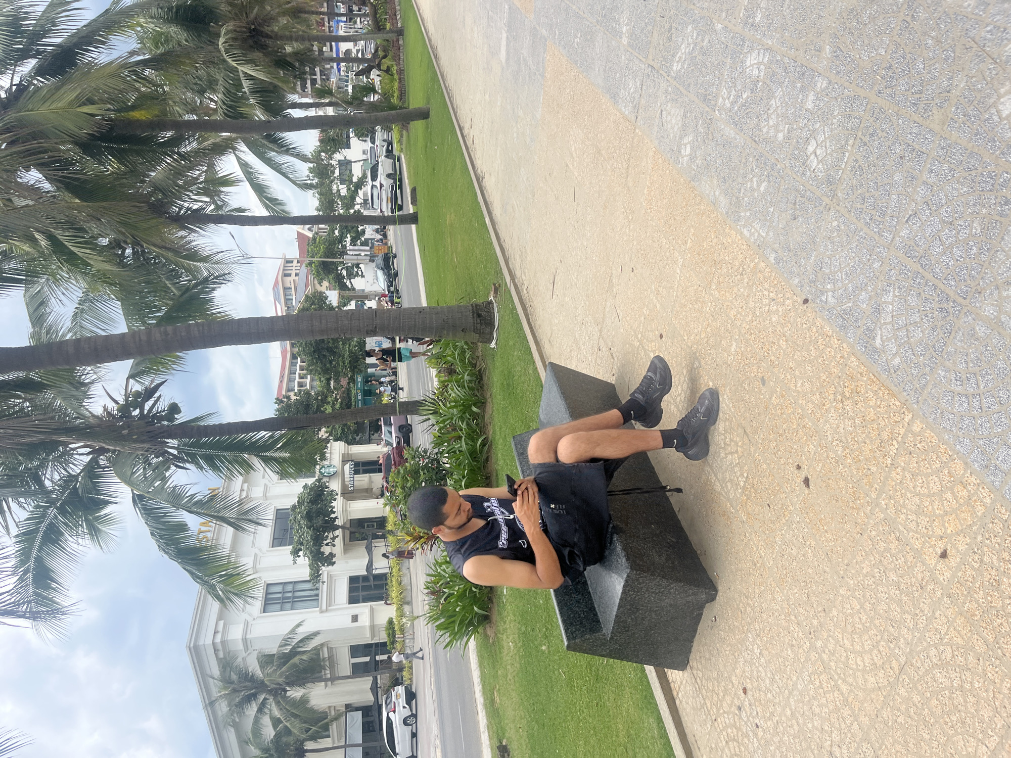 In a city scene in Da Nang, Vietnam, a man sits on a modern, dark stone bench, looking down at his smartphone. He is wearing a black sleeveless shirt with white lettering, black shorts, and black sneakers with white socks. The bench is situated on a paved walkway with a geometric pattern, bordering a grassy area with lush green grass and palm trees. Behind the grass, a street is visible with several white cars and other vehicles. Across the street, a prominent white building with classical architectural elements stands, featuring arched entrances and windows, and a sign for "STARBUCKS." Palm trees line the street and sidewalk, indicating a tropical or subtropical environment. The sky is overcast with white clouds.