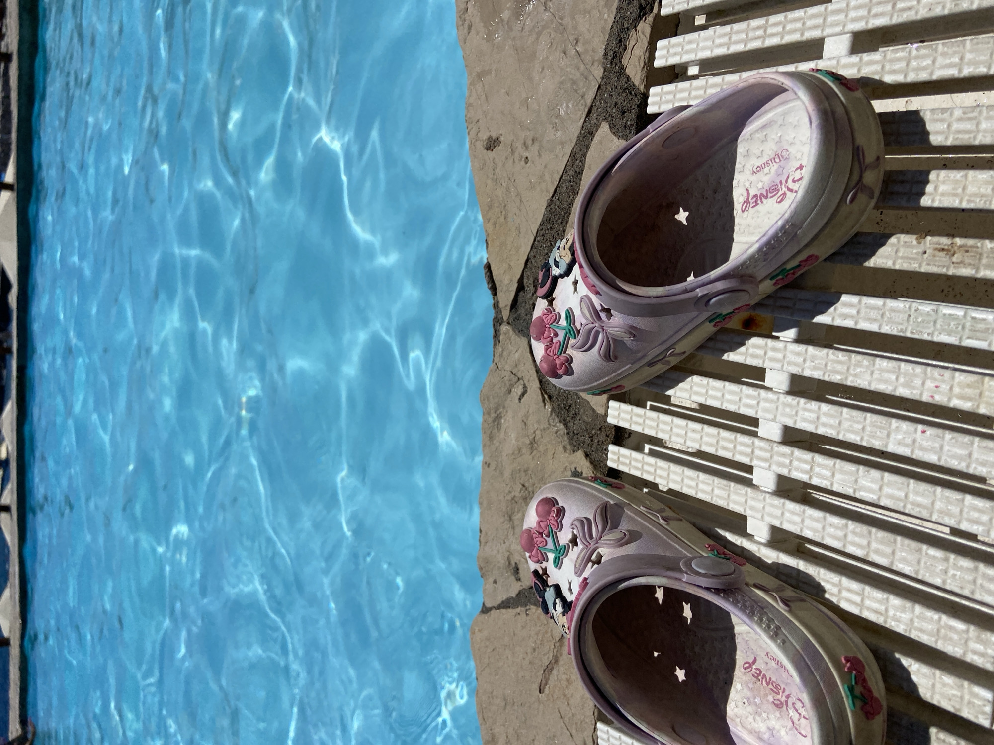 Two light purple Disney-themed clogs are placed on a white mat next to a swimming pool. The clogs are decorated with floral embellishments and character charms, possibly from Disney movies. The pool water is a vibrant blue and shows ripples and reflections from sunlight, suggesting a sunny day. The clogs are positioned on a textured, white, rectangular mat that appears to be near the edge of the pool. The ground around the pool edge is made of irregular grey stones. The image captures a moment of relaxation by the water, likely during warm weather. The scene is set outdoors, with clear lighting indicating daytime. The visible text on the inside of the clogs reads "Disney" in pink lettering. The location is identified as Prijevor, Montenegro.