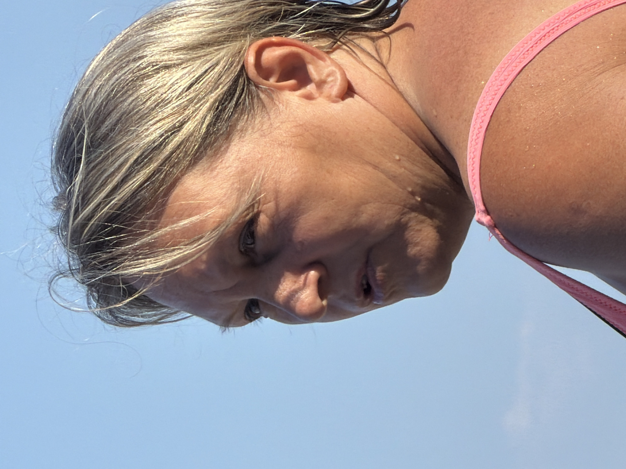 A close-up, profile shot captures a Caucasian woman with blonde, highlighted hair against a clear blue sky. Her face is turned to the left, and her mouth is slightly open. The woman appears to be wearing a pink bikini top, visible on her shoulder and chest. The lighting suggests it is daytime. The image's origin is unknown, and it is not possible to determine the location within a city.