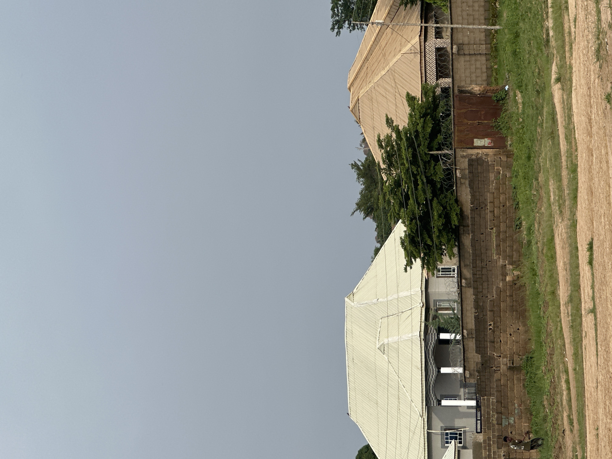 The image shows a residential area in Rukuba Road, Nigeria. In the foreground, there is a dirt path and a strip of green grass. Behind the grass is a low, rough stone wall. To the left of the wall, a young boy is walking, wearing a patterned shirt and dark pants.
Beyond the wall, two houses are visible. The house on the left has a light-colored, corrugated metal roof, a gray facade, and a porch supported by pillars. The house on the right has a reddish-brown, corrugated metal roof and a patterned wall. Both houses are partially obscured by trees. A rusty, double gate stands in the middle section of the wall, overgrown with weeds. The sky above is a uniform gray. Barbed wire is visible along the top of the wall in front of the houses.