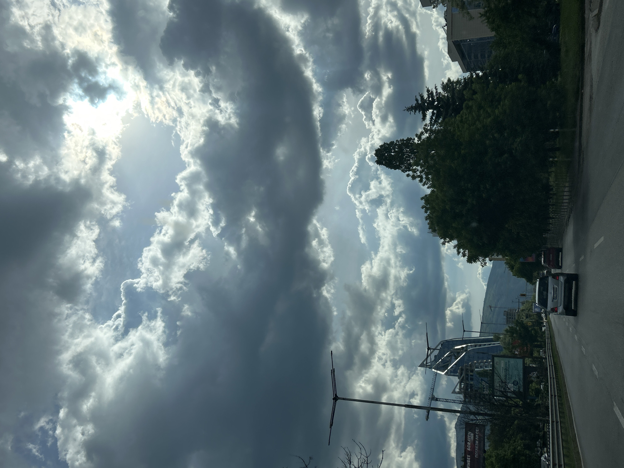 The image shows a street scene in Sofia, Bulgaria. A road runs along the right side, with two cars visible, a white van and a dark-colored car. The background includes a mountain range and construction cranes amidst buildings. A prominent building complex with reflective surfaces is partially visible, along with a large billboard. In the sky, there are large, dramatic cloud formations, with sunlight breaking through. The clouds dominate the upper portion of the image. Power poles are visible along the roadside.