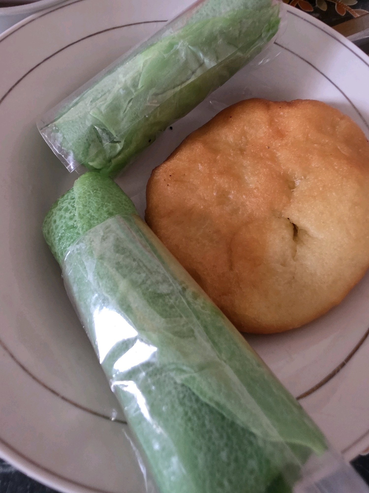 A close-up shot shows a white plate with a thin brown rim. On the plate are two green, rolled snacks individually wrapped in clear plastic and one golden-brown, fried pastry. The green snacks appear to be made of a delicate, crepe-like material, possibly filled with something. The pastry is round and slightly puffed, with some indentations on its surface. The background is slightly blurred, showing a darker patterned surface. The scene is likely from a kitchen or dining area in Tanjung Selor Timur, Indonesia, given the typical Indonesian snacks depicted.