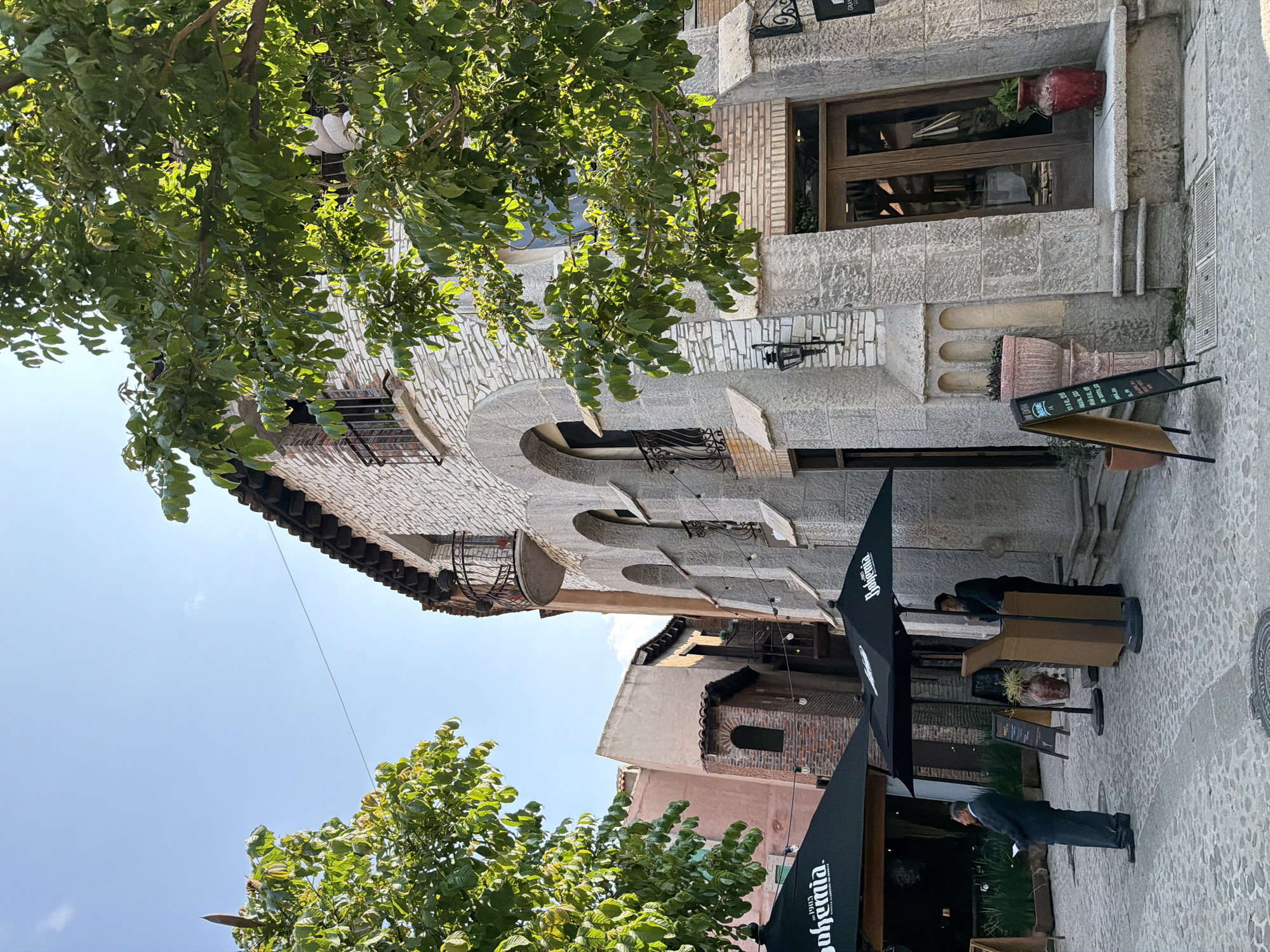 An outdoor shot shows a two-story building constructed from stone and brick, featuring arched windows with wrought-iron balconies. An open window box and decorative stone elements accentuate the façade. A doorway, framed by stone, leads into the building. Outdoor seating with umbrellas is set up on the cobblestone street. Two individuals wearing suits are observed, one standing and the other seated. Above, tree foliage partially obscures the building. The shot is taken in Domingo Arenas, Mexico.