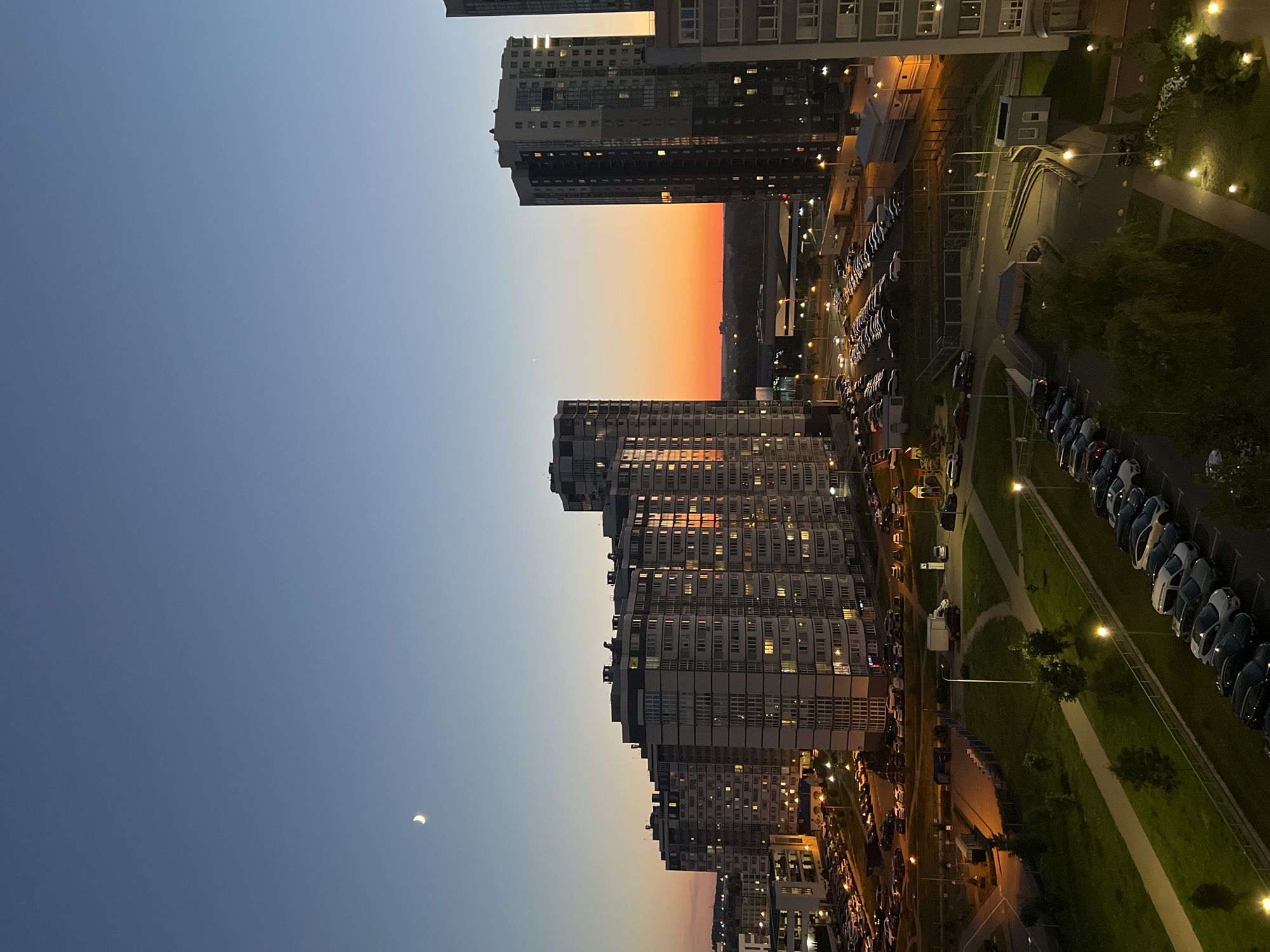This vertical, eye-level, medium shot captures a cityscape at dusk, featuring modern high-rise apartment buildings and a vibrant sunset sky. The sky transitions from a deep blue at the top to a bright orange and yellow band just above the horizon, with a sliver of the moon visible in the upper left quadrant. A row of tall, multi-story residential buildings dominates the middle ground, their windows illuminated, casting a warm glow. In the foreground, there is a parking lot filled with numerous cars, and a section of a green park with lampposts. The scene suggests a residential district within the city of Minsk, Belarus, given the architectural style of the buildings. There are no discernible people or animals in the frame. The overall impression is one of urban tranquility at the close of day.
