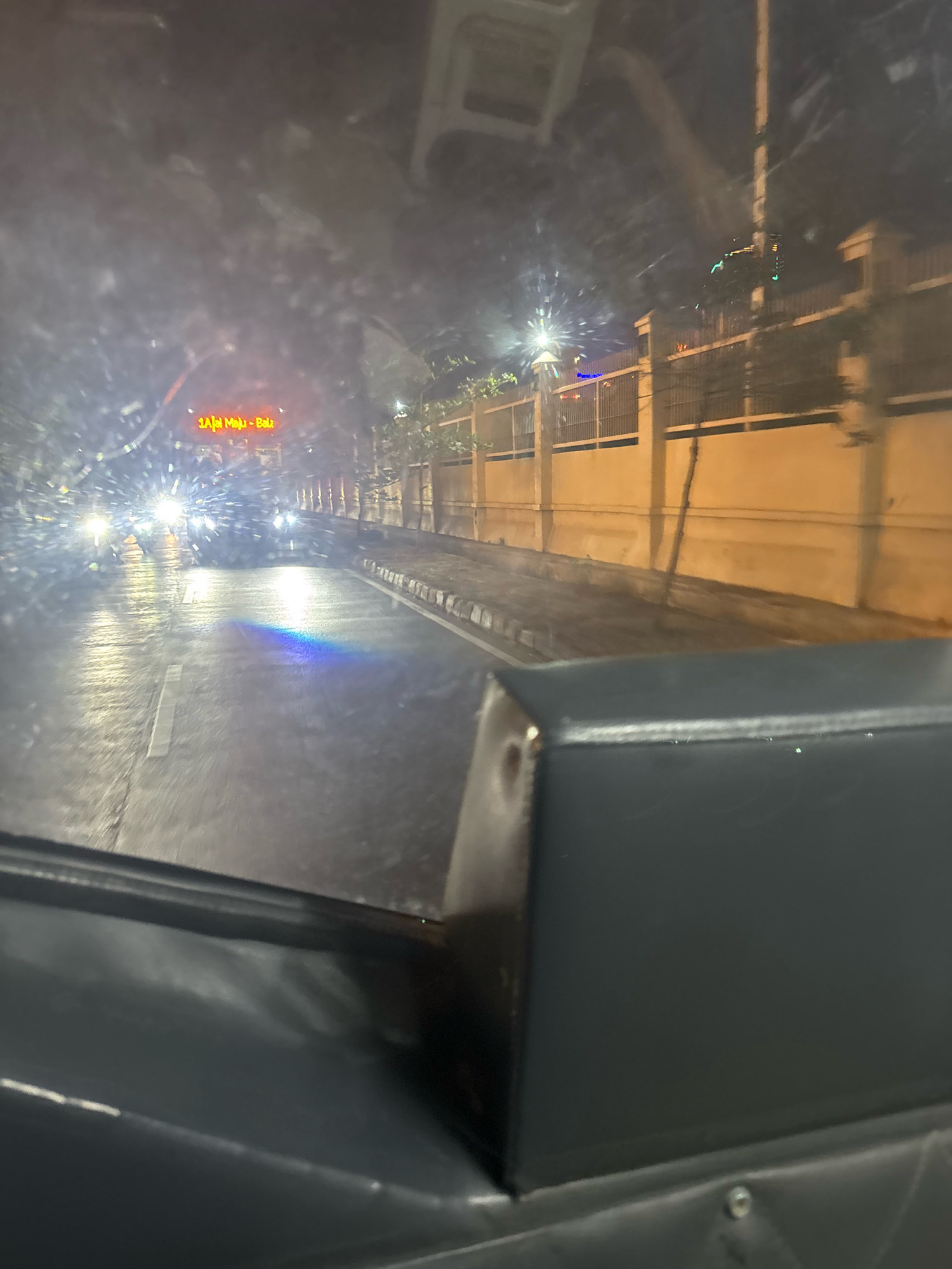 The image shows a view from inside a vehicle at night, looking out onto a wet street. The vehicle's dashboard is visible in the foreground, appearing dark gray. Outside, traffic lights and streetlights illuminate the scene. Several vehicles are on the road, with their headlights on, suggesting it might be raining or the road surface is wet. There is a fence with lights along the right side of the street, and a building behind it. A neon sign with red text is visible above the approaching vehicles. Based on the street signs and architectural style, this scene is likely located in North Jakarta, Indonesia. The overall impression is a typical urban night scene with vehicle traffic and street lighting.