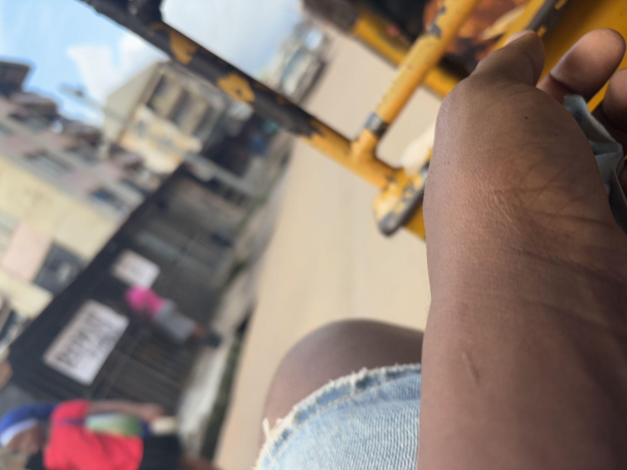 The image depicts a close-up view of a person's hand and leg, wearing denim shorts, positioned within what appears to be a yellow tricycle or a similar open-air vehicle. The hand is resting on the leg, and a folded piece of paper or bill is visible near the fingers. In the background, the scene is blurred, but it suggests an urban environment in Onitsha, Nigeria, characterized by buildings and a street. Two figures are visible in the mid-ground: one wearing a red shirt and a blue head covering, and another in a pink shirt. The overall impression is of a candid, everyday moment captured while in transit.