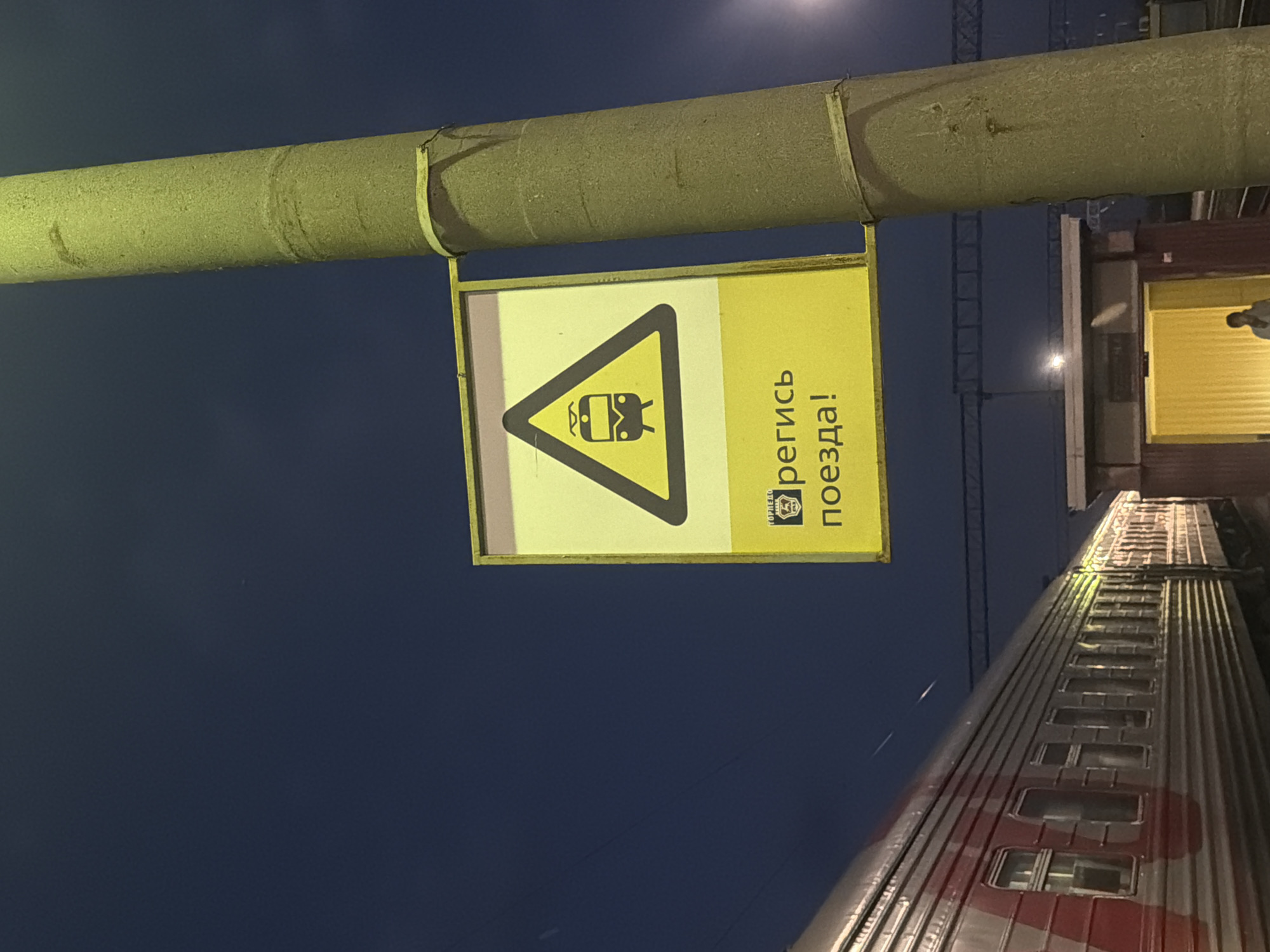 The image depicts a nighttime scene at a train station. A yellow warning sign with a black border is attached to a concrete pole on the right side of the frame. The sign contains a triangular symbol of a train and the Russian text "Берегись поезда!" which translates to "Beware of the train!". To the left of the pole, a long, silver train with red accents is parked. The train cars have multiple windows. In the background, there is a building with a brightly lit entrance and a person standing within it. Overhead power lines are visible above the train. The sky is dark blue, indicating it is night. The overall setting suggests a train platform or station area. The location is identified as the city of Balashov, Russia.