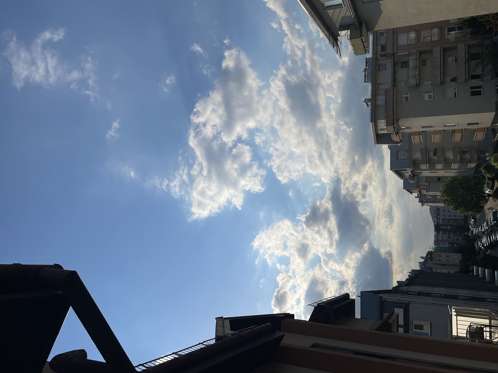 The image shows a view of a city street from a low angle, looking upwards. The sky is predominantly blue, with scattered white cumulus clouds. Buildings are visible on both sides of the frame, with multiple windows and balconies. There are cars parked along the street. In the foreground, a dark roof of a building is visible, creating a silhouetted effect. The location is in an urban area with residential buildings.
