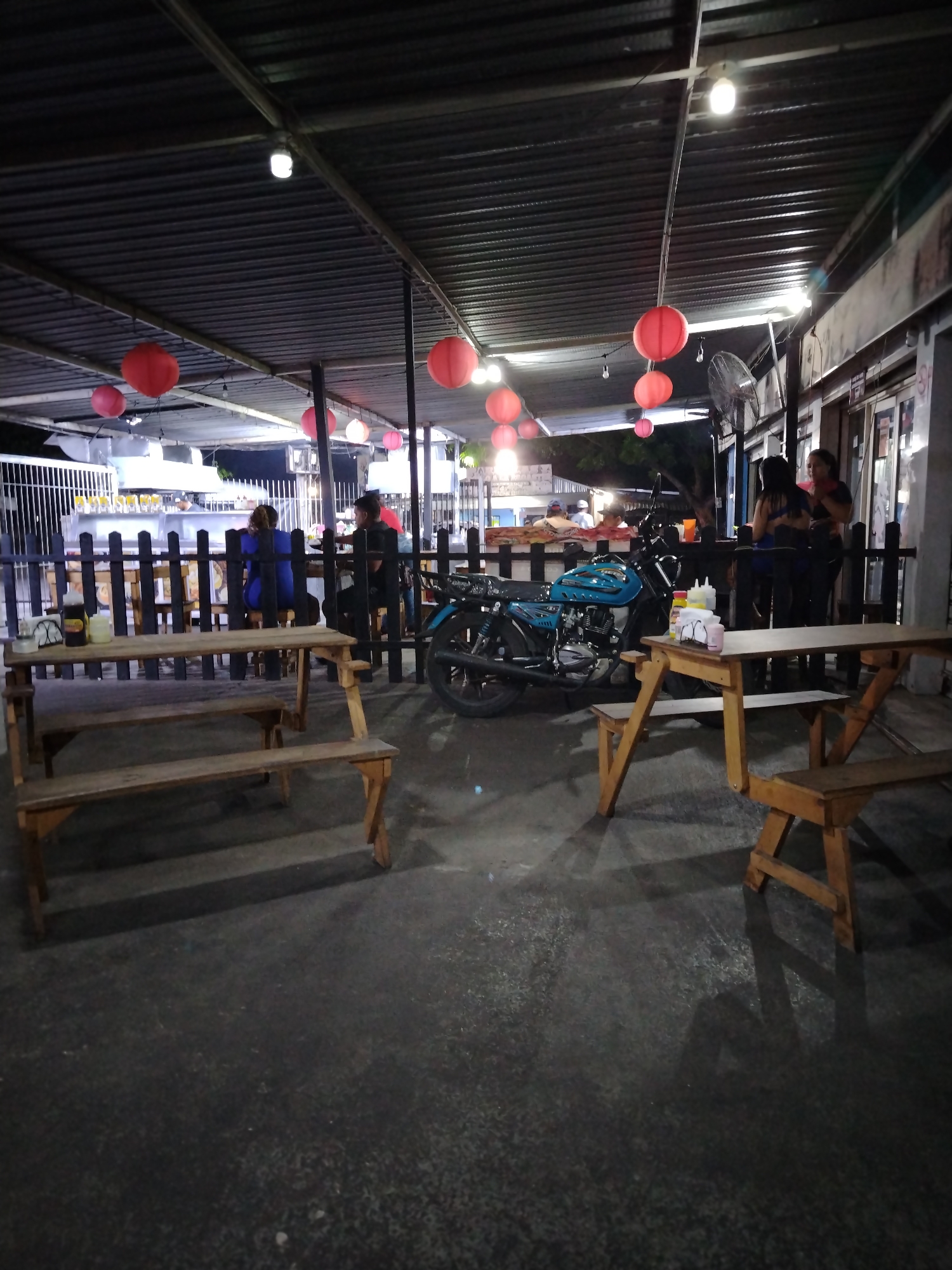 The image shows an outdoor eating area in Tinaquillo, Venezuela, at night. The space is covered by a corrugated metal roof with hanging red lanterns and light bulbs. People are seated at picnic tables behind a dark wooden fence, while others stand near the edge of the covered area. A blue motorcycle is parked between the tables. Picnic tables are in the foreground, one with bottles on it. The ground is a dark, paved surface.