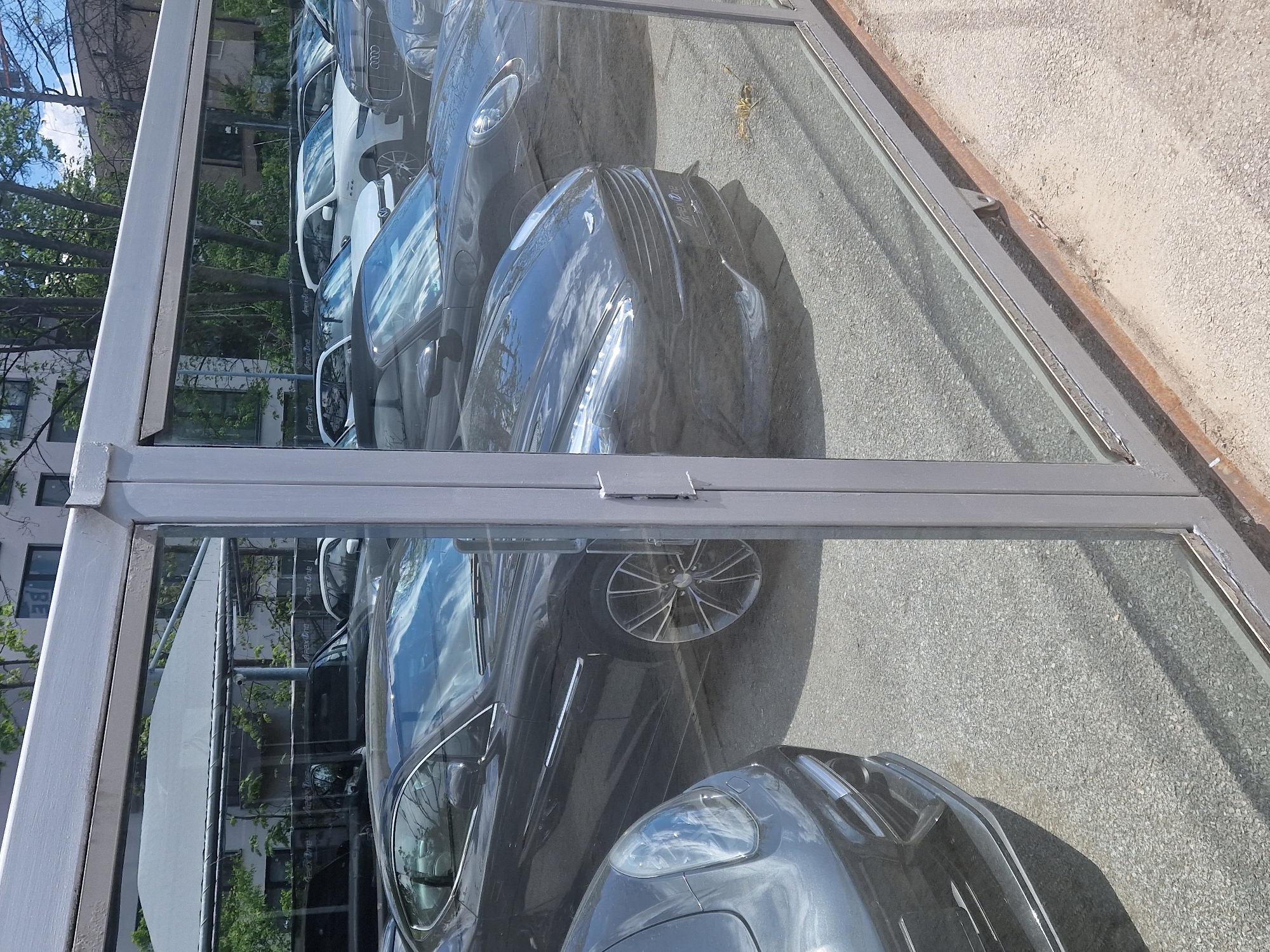 The image shows a car dealership's display of cars through a window, likely in a European urban environment. The vehicles, including at least one Aston Martin and several Audis, are parked closely together. The reflections in the window show trees and a building across the street, indicating an urban setting. The dealership appears to be modern, with glass and metal structural elements. No people are directly visible. The focus of the scene is on showcasing the cars, and the environment suggests a business district.