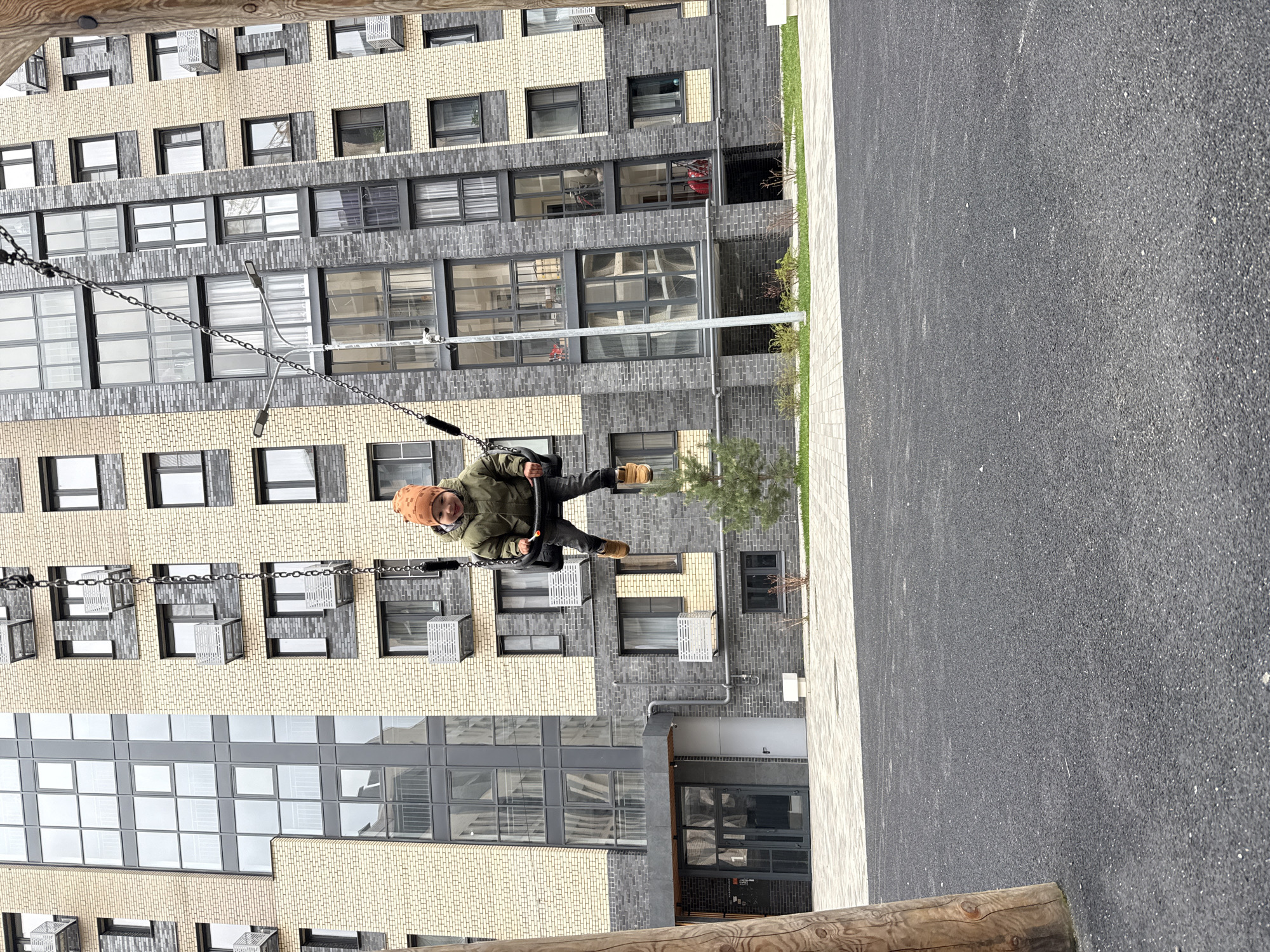 A young child in a brown hat and green jacket is on a swing in front of a large, modern apartment building. The building has a facade of alternating light beige and dark gray bricks, with many windows. The ground beneath the swing is a dark gray, textured material. A wooden beam is visible in the foreground at the bottom of the image. The photograph appears to be taken at ground level.
The city mentioned, Rusanovo, Russia, is simply an added detail and there is no verification this photo was taken there.