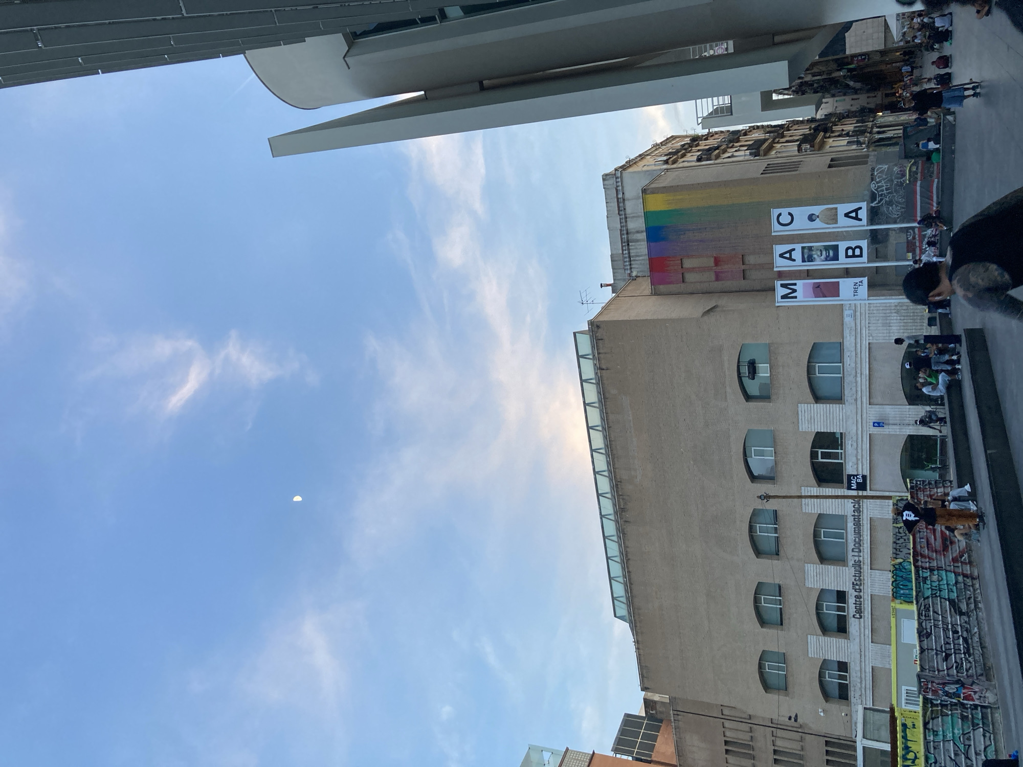 The image shows a low-angle view of a street scene in Barcelona, Spain, likely in the El Raval district, a vibrant area known for its cultural institutions and street art. The sky is a bright blue with scattered white clouds and a visible crescent moon. Dominating the center of the frame is a large, light-brown brick building with arched windows. A rainbow-colored mural is painted on the side of this building, adding a splash of color to the facade. Three tall banners with the letters "M A C B A" are displayed in front of the building.
To the right of this building, a modern, white structure with sharp geometric lines rises upwards, contrasting with the older architecture. In the foreground, a dark gray paved area serves as a public space. Several people are present, including individuals sitting and standing. Some are gathered near a low wall covered in graffiti, while others are spread out on the ground. One person in the foreground is standing and appears to be interacting with others. Another person in the mid-ground is sitting on a step, wearing a black tank top and shorts, with a skateboard nearby. The overall atmosphere suggests a public gathering or an outdoor event. The "Centre d'Estudis i Documentació MACBA" is visible on the building, identifying it as part of the Museu d'Art Contemporani de Barcelona (MACBA).