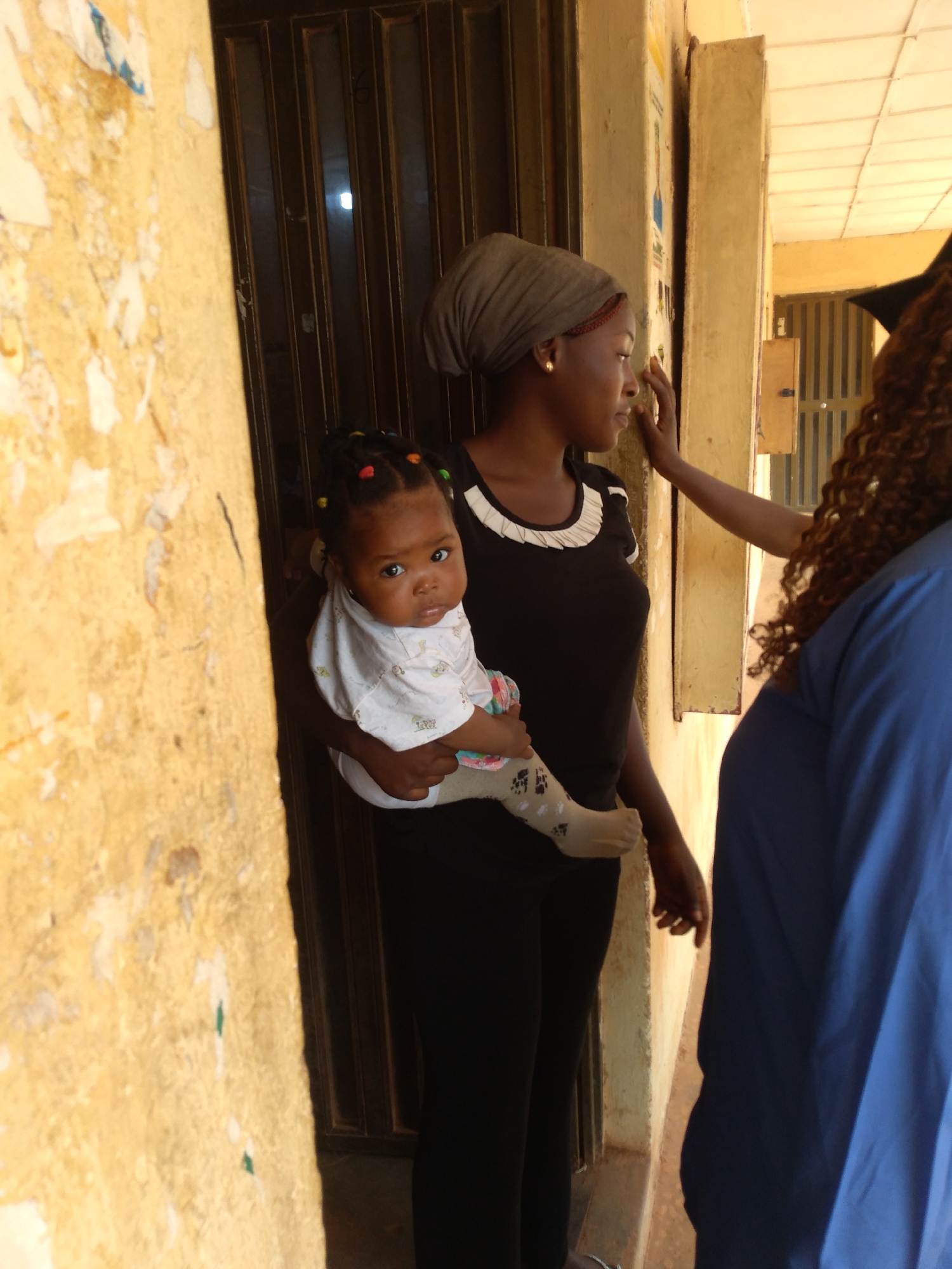 A young Black woman wearing a grey headscarf and a black shirt is standing outside a building. She is holding a baby wearing a white shirt with a pattern of small green and blue drawings. The baby has multi-colored beads in its hair. Another Black woman, whose back is to the camera, is wearing a blue garment. The scene appears to be in a city location, possibly in Jos, Nigeria, given the style of the building's facade and the presence of posters on the wall. The wall to the left is peeling and discolored, with visible patches and tears. The background shows a dark doorway or entrance with vertical metal bars. The lighting suggests it is daytime.