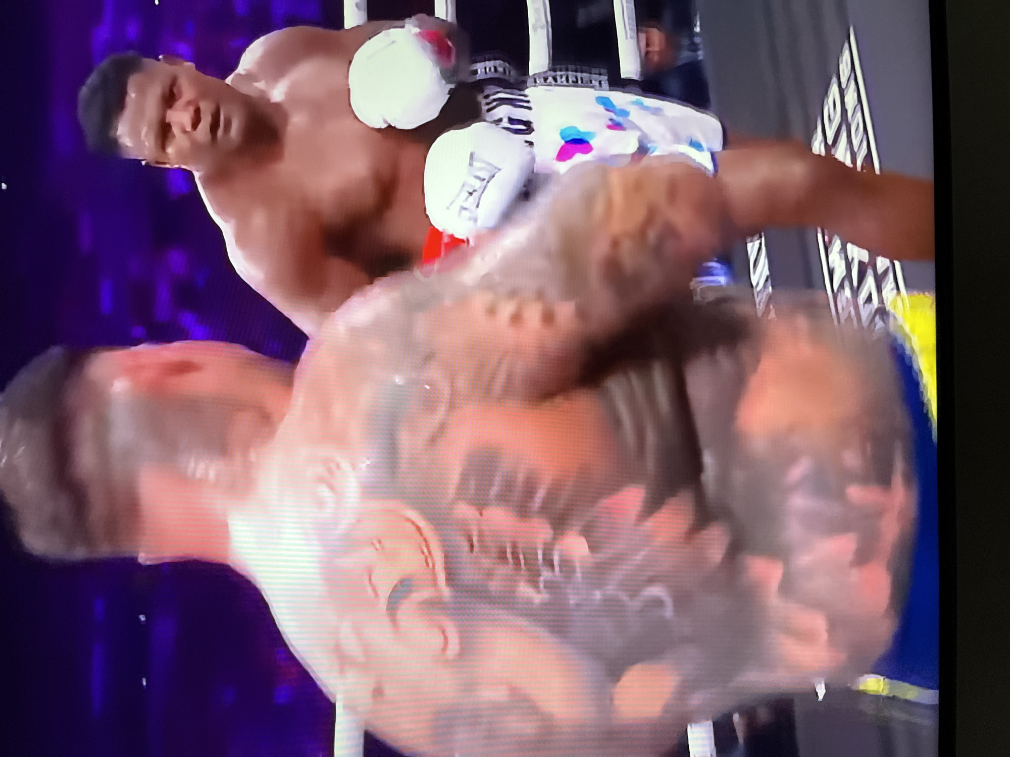 Two men in boxing shorts and gloves are in a boxing ring with a purple, blurred background. The man on the left has a large tattoo on his back and is facing away from the camera. The man on the right is facing the camera with a neutral expression and is wearing white boxing gloves. He is wearing white shorts with colorful butterfly designs and black stripes. The boxing ring has white ropes and a black floor with white lettering. There are spectators in the background, also blurred. The scene appears to be a live boxing match, possibly in a stadium or arena. There is no visible information to indicate the location in Kharkiv, Ukraine.