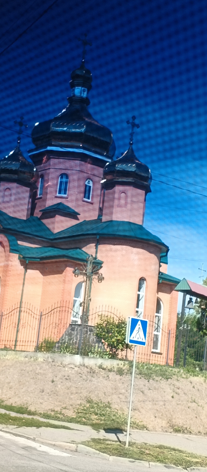 The image displays the exterior of a brick church with several domes, featuring green roofs and crosses on top. The church is situated in an urban environment with a blue sky visible in the background. In the foreground, a road with a pedestrian crossing is visible, alongside a traffic sign indicating a pedestrian crossing. A metal fence borders the church property. The image appears to be taken from a slightly elevated perspective, possibly from a moving vehicle. There are no people or visible actions depicted in the scene. The overall impression is of a religious building in an accessible urban area.