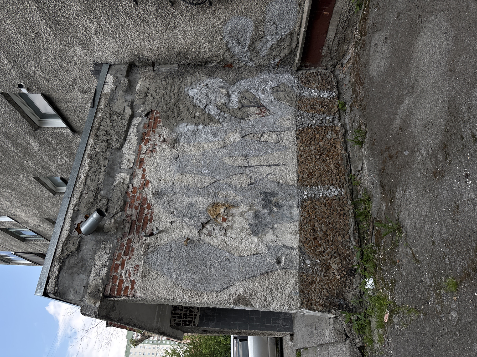 This is a photograph of the exterior of an aged, grey concrete apartment building located in Novosibirsk, Russia. The building features multiple windows with white frames and a textured, weathered facade. In the foreground, a section of the building's lower wall is adorned with a crude, faded mural or graffiti depicting abstract human-like figures in shades of grey. Below the mural, the wall is covered in a band of small, rounded brown and grey stones. To the left of the mural, a doorway is visible, partially obscured by a metal grate. A downspout extends from the roofline over the muraled section. The ground in front of the building is a rough, dark asphalt surface with patches of dirt and weeds. Some of the asphalt appears wet. The sky above is overcast with some clouds visible. In the background, other apartment buildings and trees can be seen.