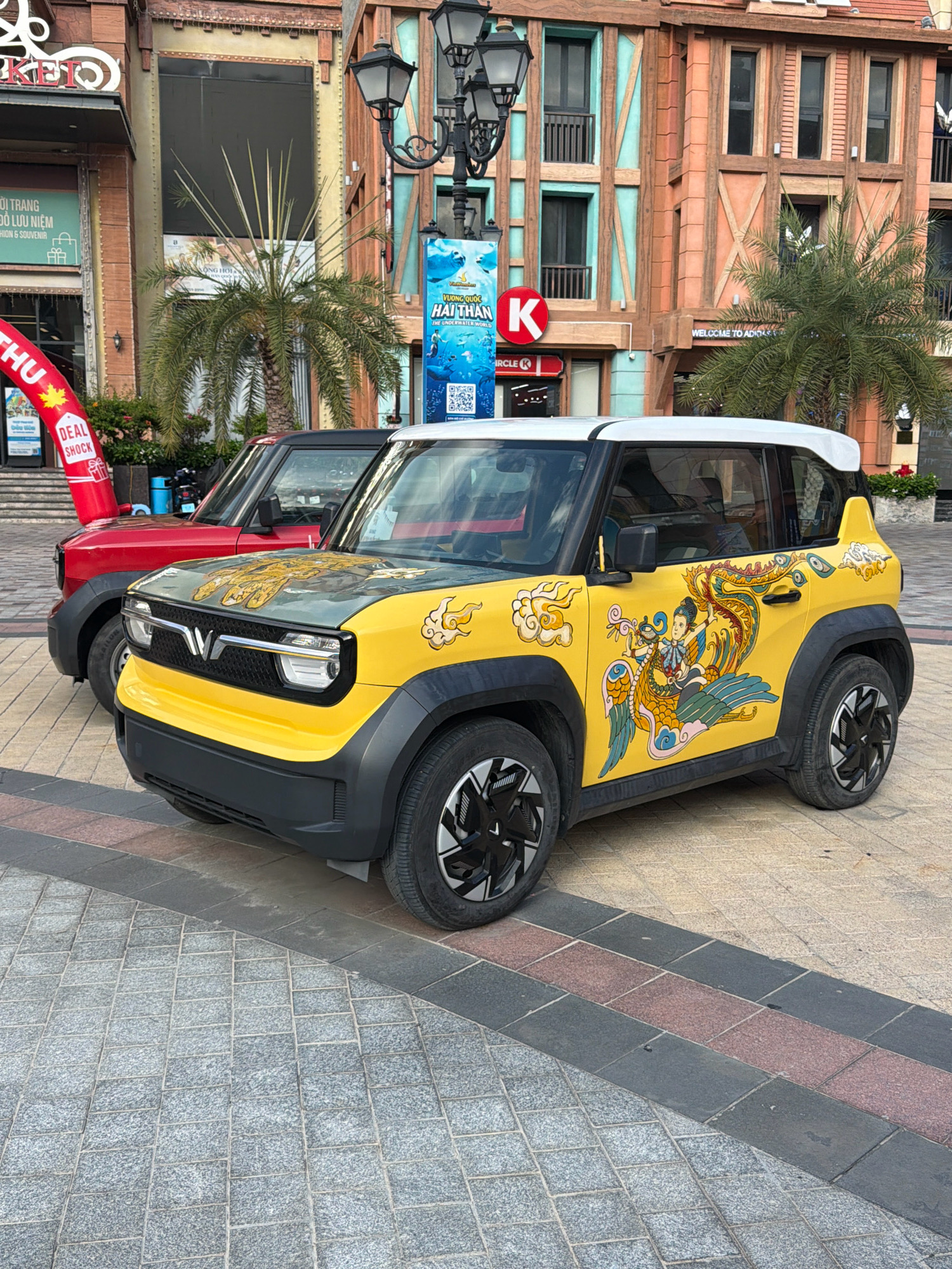 The image depicts two compact electric cars parked on a paved walkway in Nha Trang, Vietnam. The car in the foreground is yellow with a white roof and features a prominent, colorful decal on its side depicting a traditional dragon and a figure in an ornate costume. The decal on the hood appears to be a similar style. The car behind it is red. The backdrop includes a building with a facade resembling European architecture, featuring colorful panels, wooden beams, and black-framed windows. A tall, ornate lamppost stands to the left of the building. Various signs are visible, including one for "Circle K" and another with Vietnamese text and graphics of marine life. Palm trees are interspersed throughout the scene, suggesting a warm climate. The walkway is paved with interlocking grey and reddish-brown square stones, with darker grey rectangular stones forming a border. The overall scene suggests a commercial or recreational area within the city.