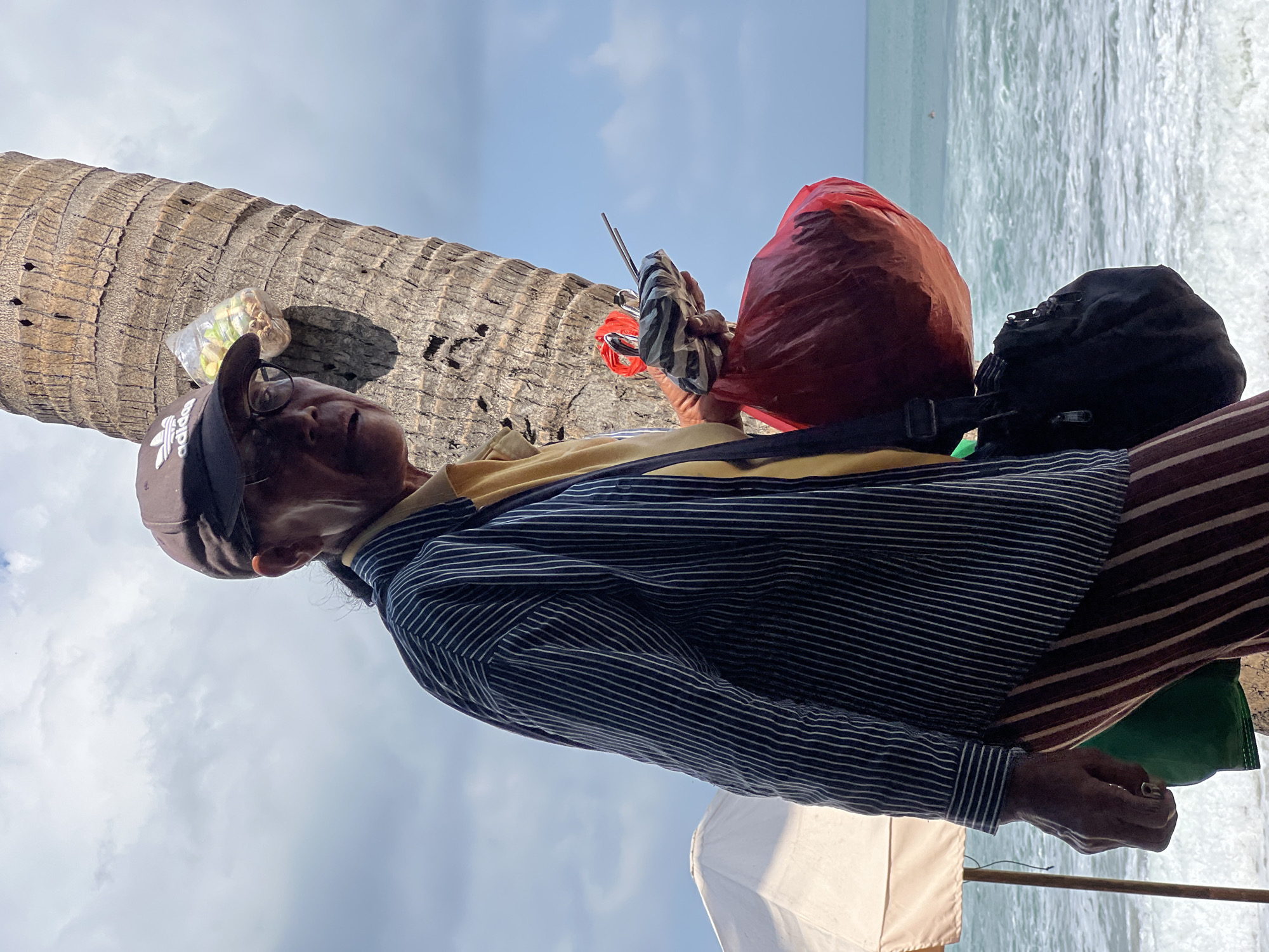 A man in a brown Adidas baseball cap and glasses stands by a palm tree on a beach in Kuta, Indonesia. He wears a yellow shirt under a dark blue and white pinstriped button-down shirt and striped pants. He holds a red plastic bag and a folded striped object in his hands. A black shoulder bag is slung across his body. A white beach umbrella is visible in the background, along with the ocean and a cloudy sky. A clear plastic bag containing snacks is attached to the palm tree.