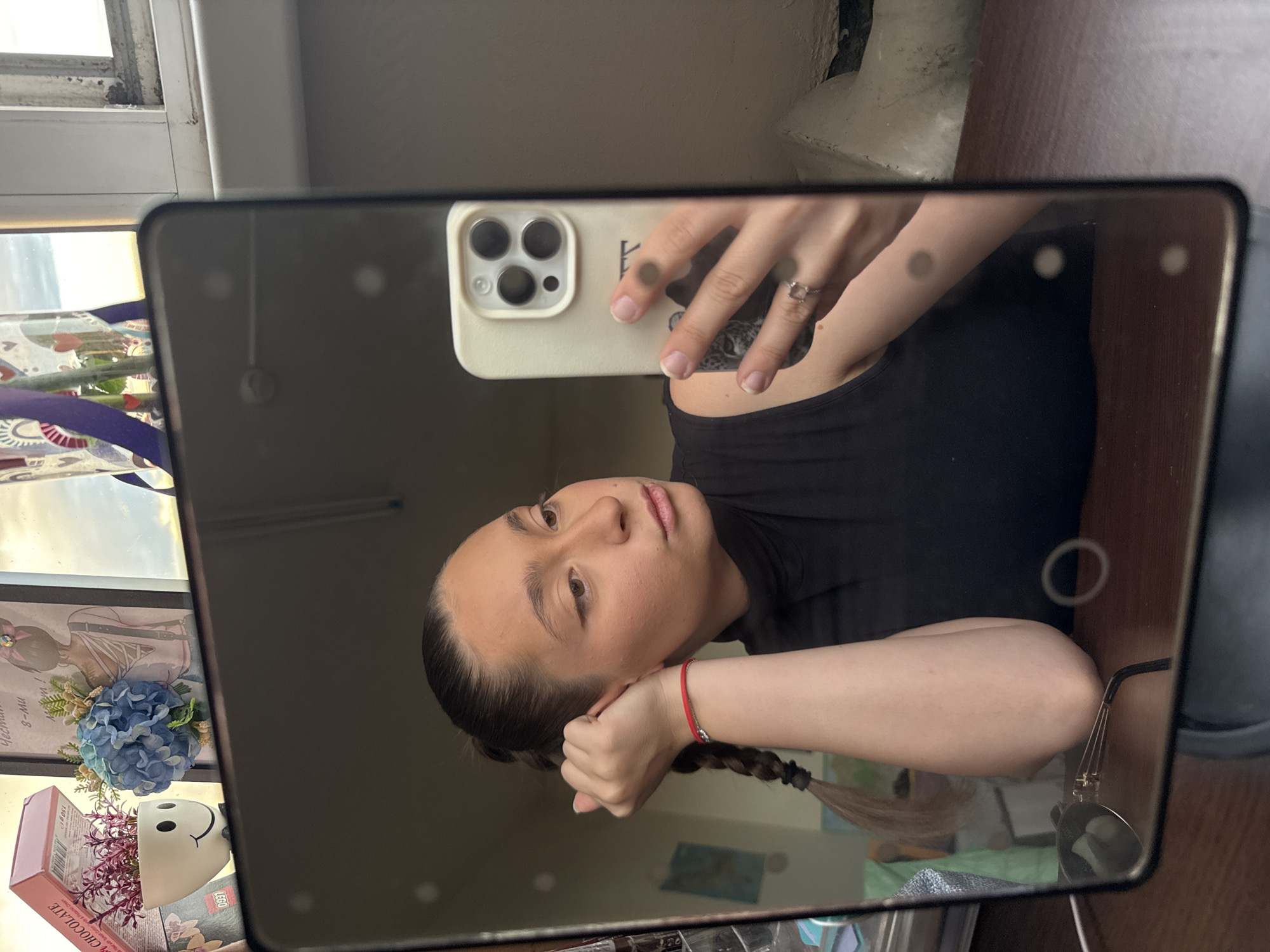 A young woman with brown hair in a braid, wearing a black sleeveless top and a red bracelet, is taking a selfie in a mirror. She is resting her head on her hand, looking away from the camera. A smartphone with a light-colored case is visible in her hand. The mirror reflects items on a shelf behind her, including a box of chocolates, a white pot with a smiley face and purple plants, and a framed picture of a woman with a blue flower. The environment appears to be indoors, possibly a bedroom or dressing area, with a window visible in the background. The lighting suggests it is daytime. The background also shows a wooden desk surface and some dust on the window frame. No specific landmarks indicating Sofia, Bulgaria, are visible.