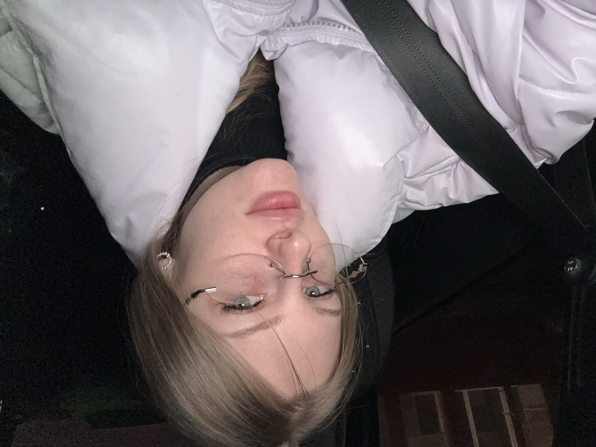 A young person is angled in a seated position in what appears to be a vehicle, wearing a light purple puffer jacket and a black seatbelt. The person has blonde hair, light skin, and is wearing rimless glasses. The background is mostly dark, suggesting an interior setting, possibly within a car. The specific location in Perm, Russia, is not discernible from the image.