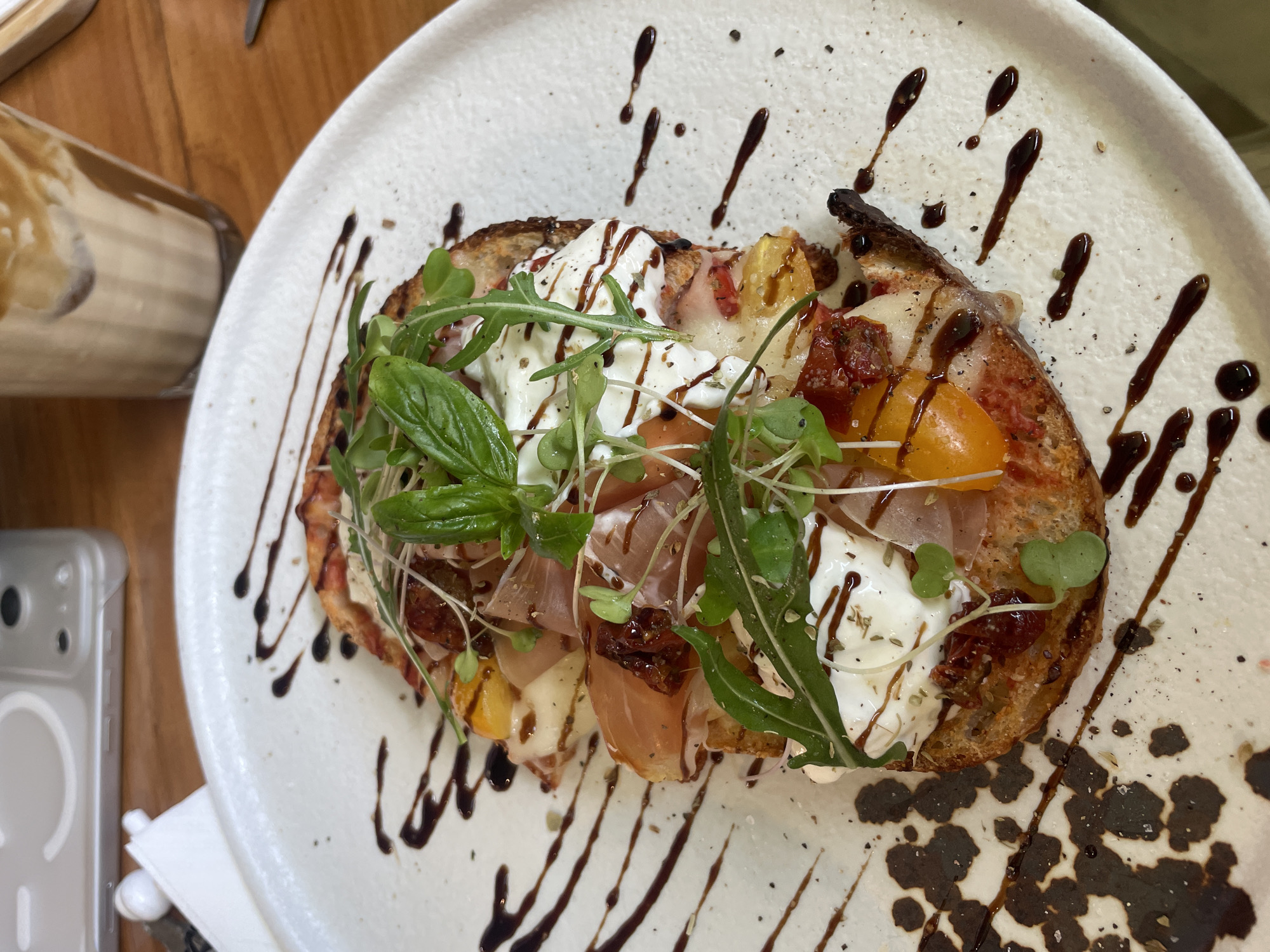 A plate of food sits on a wooden table. The plate is white and speckled with gray, adorned with a brown balsamic glaze. The food item is a toasted slice of bread topped with cheese, basil, sprouts, tomatoes, and prosciutto. A glass of liquid with layers is partially visible to the left of the plate, along with a phone. The photograph was likely taken indoors. The image is not clearly indicative of the location being Choeng Thale, Thailand.