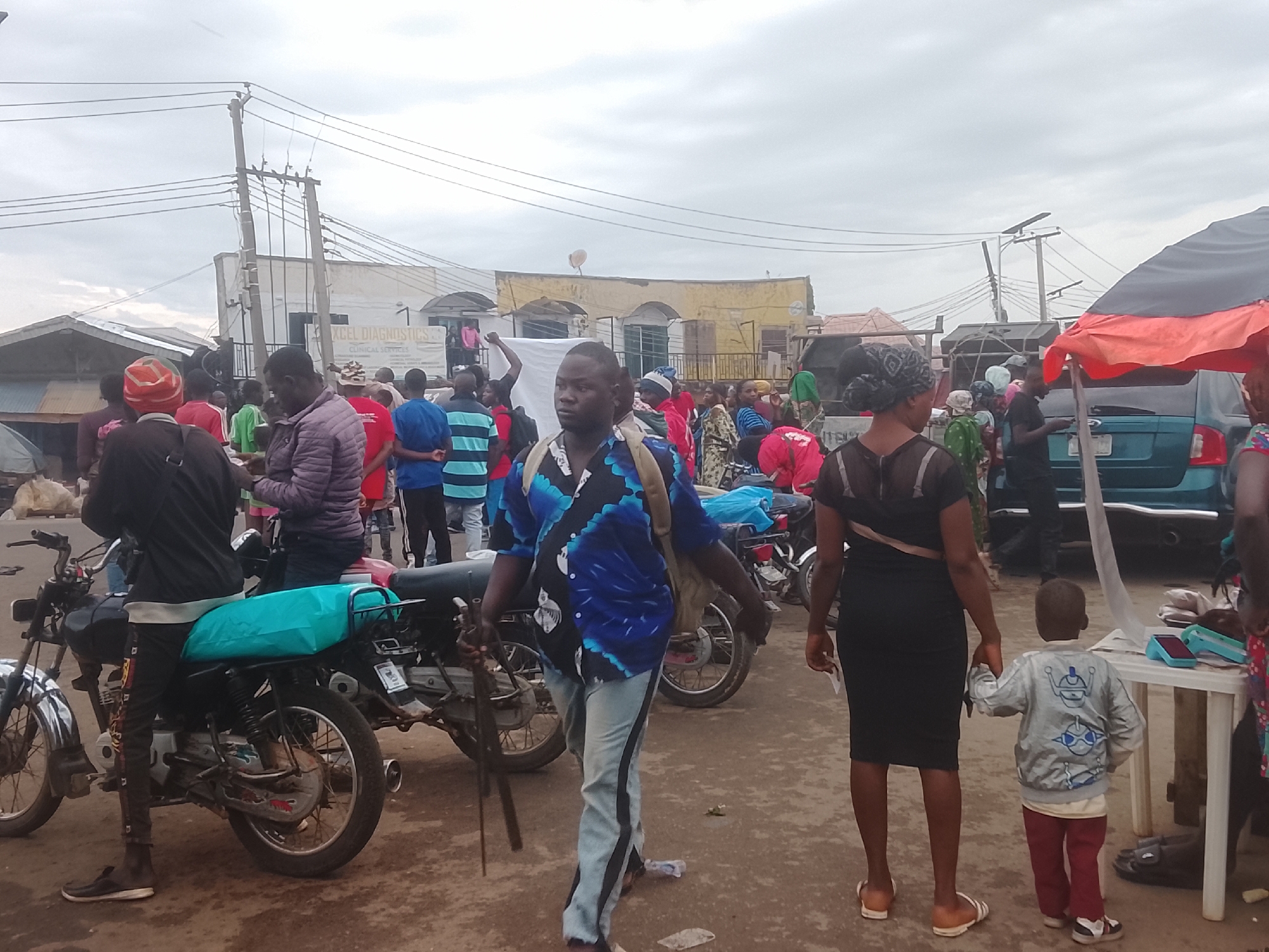 A street in Gwong, Nigeria is crowded with people. Several motorcycles are parked. A man in a blue patterned shirt and light jeans holds two dark sticks. A woman in a black dress and sandals holds the hand of a child wearing a gray jacket and red pants. A teal minivan is visible. Buildings line the background, and electrical wires hang overhead. The sky is overcast.