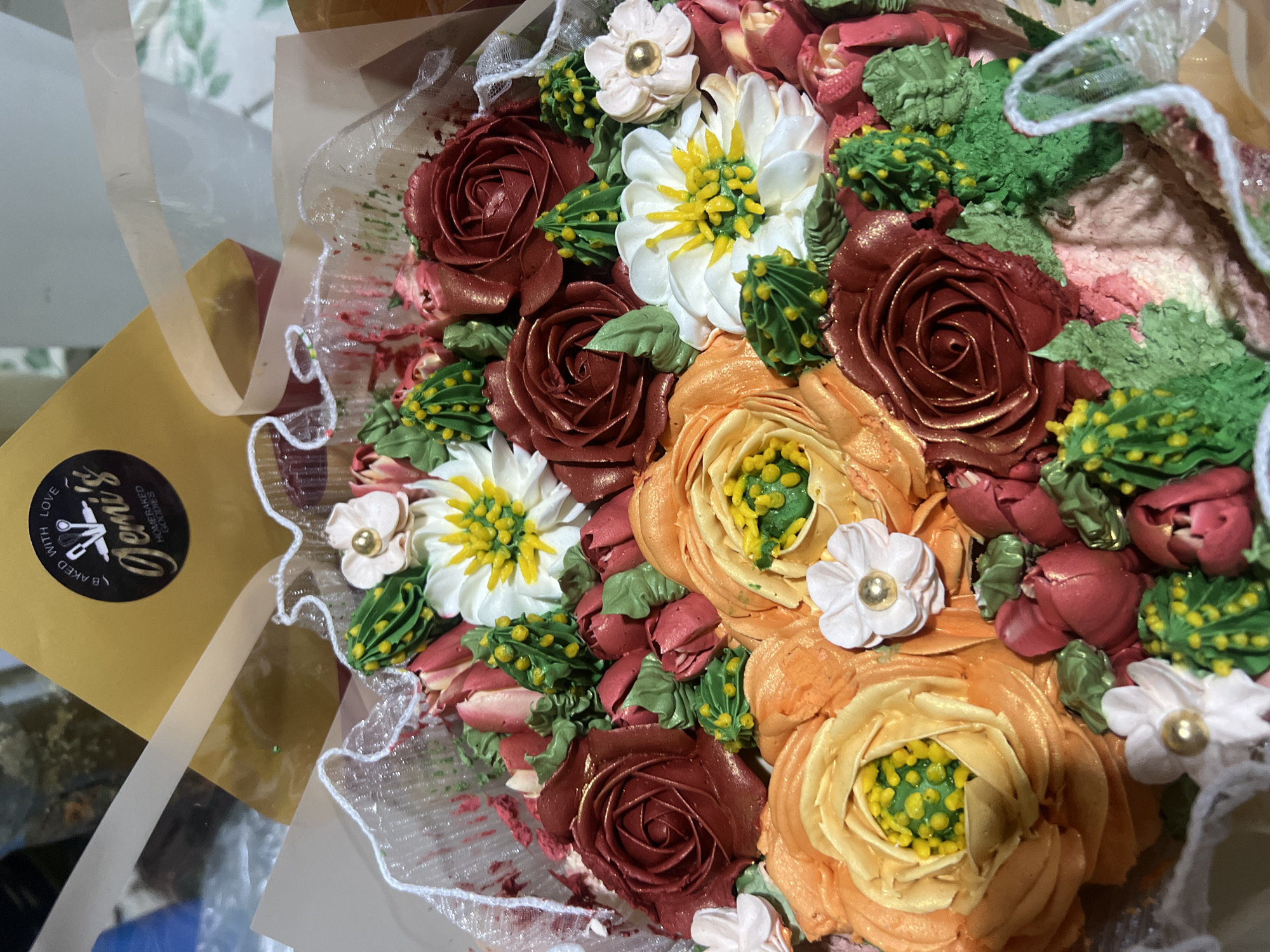 The image shows a close-up, overhead view of a decorated cake. The cake is adorned with an arrangement of buttercream flowers in various colors, including deep red roses, orange and yellow flowers, white daisies with yellow centers, and small pink buds. Green buttercream leaves and cactus-like accents are interspersed among the flowers. The cake appears to be set in a container, with decorative white lace trim visible around the edge. In the upper background, a golden sticker with black text reads "Jemi's HOMERBAKED GOODIES." The setting is indoors, with a blurred background that suggests a domestic or commercial environment, but no specific urban or natural features are discernible. The image does not provide any information about the city of Panabo, Philippines.