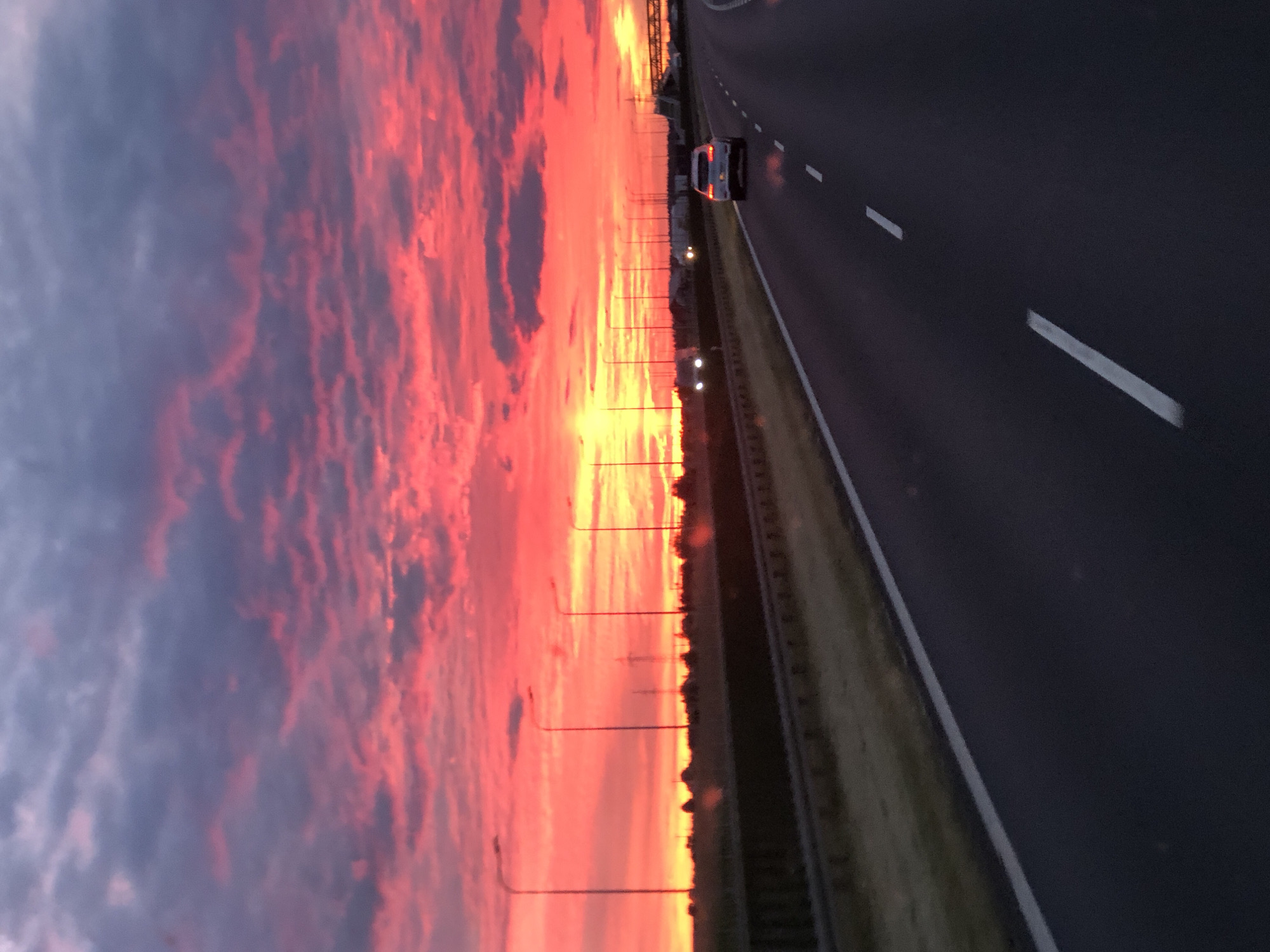 The image depicts a highway scene at sunset in Zaparcin, Poland. The sky is dominated by vibrant hues of pink and orange, with clouds casting dramatic shadows. A white SUV is driving on the right lane of the highway, while other vehicles are visible in the distance. Streetlights line the side of the road, and a guardrail separates the highway from the adjacent landscape. The overall impression is one of a scenic drive during a picturesque sunset.
