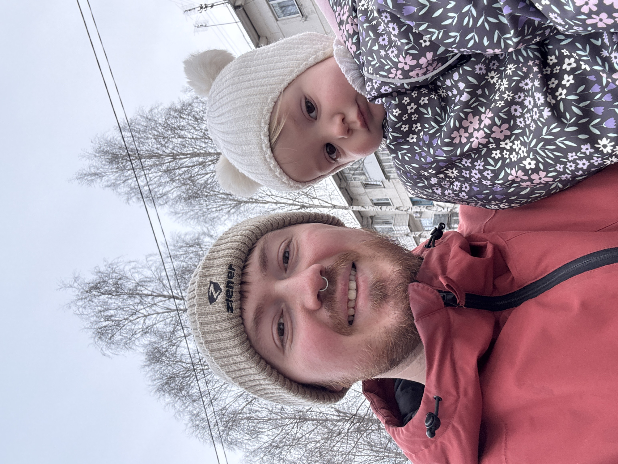 A selfie photograph captures a fair-skinned man and a young child. The man is wearing a tan knit beanie with a "Ziener" logo and a red jacket. He has a nose ring and a beard. The child is on his shoulders, wearing a white pom-pom hat and a floral jacket. The background features bare trees, overhead utility lines, and a multi-story building, indicating an urban or suburban setting. The location is identified as Chaikovsky, Russia, based on location data. The orientation suggests the photo was taken with the camera held above the subjects.