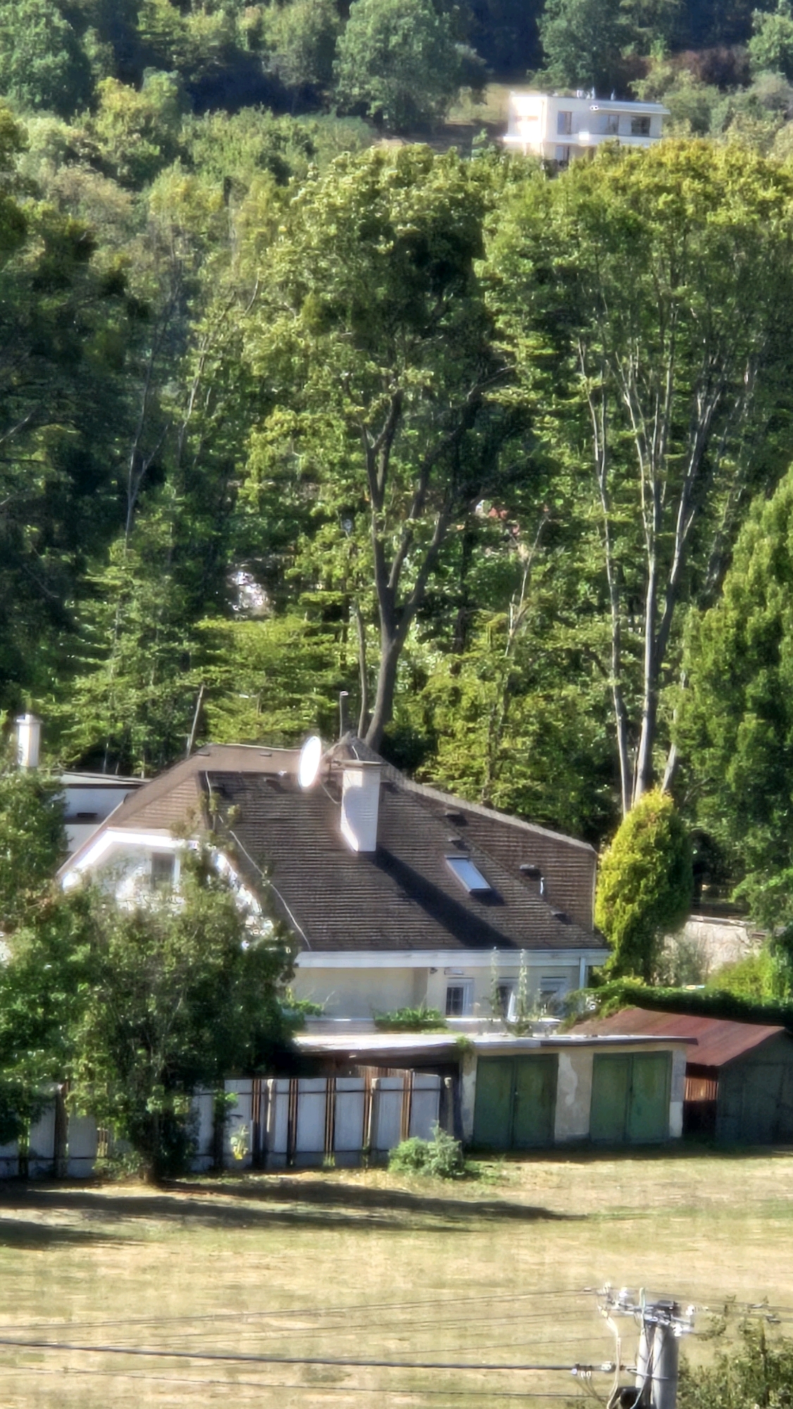 The image depicts a residential area nestled among lush green trees and foliage. In the foreground, a dry, grassy field with thin, horizontal wires suggests a rural or semi-rural setting. A gray electrical pole with wires is visible in the lower right corner.
In the midground, a modest house with a dark, sloped roof is partially obscured by trees. The house features a white chimney and a satellite dish. Adjacent to the house is a covered structure with a slatted wooden fence and two green garage doors. To the right of this structure, a smaller, dark-colored shed with a red roof is visible. The area around the buildings appears somewhat overgrown with vegetation.
In the background, the dense canopy of trees dominates the landscape. Higher up on a hill, a modern, light-colored house with multiple windows is visible, bathed in sunlight. The overall impression is one of a quiet, suburban or semi-rural environment with a mix of older and newer residential buildings surrounded by nature. The location is identified as being in the city of Prešov, Slovakia. There are no people or animals visible in the image.