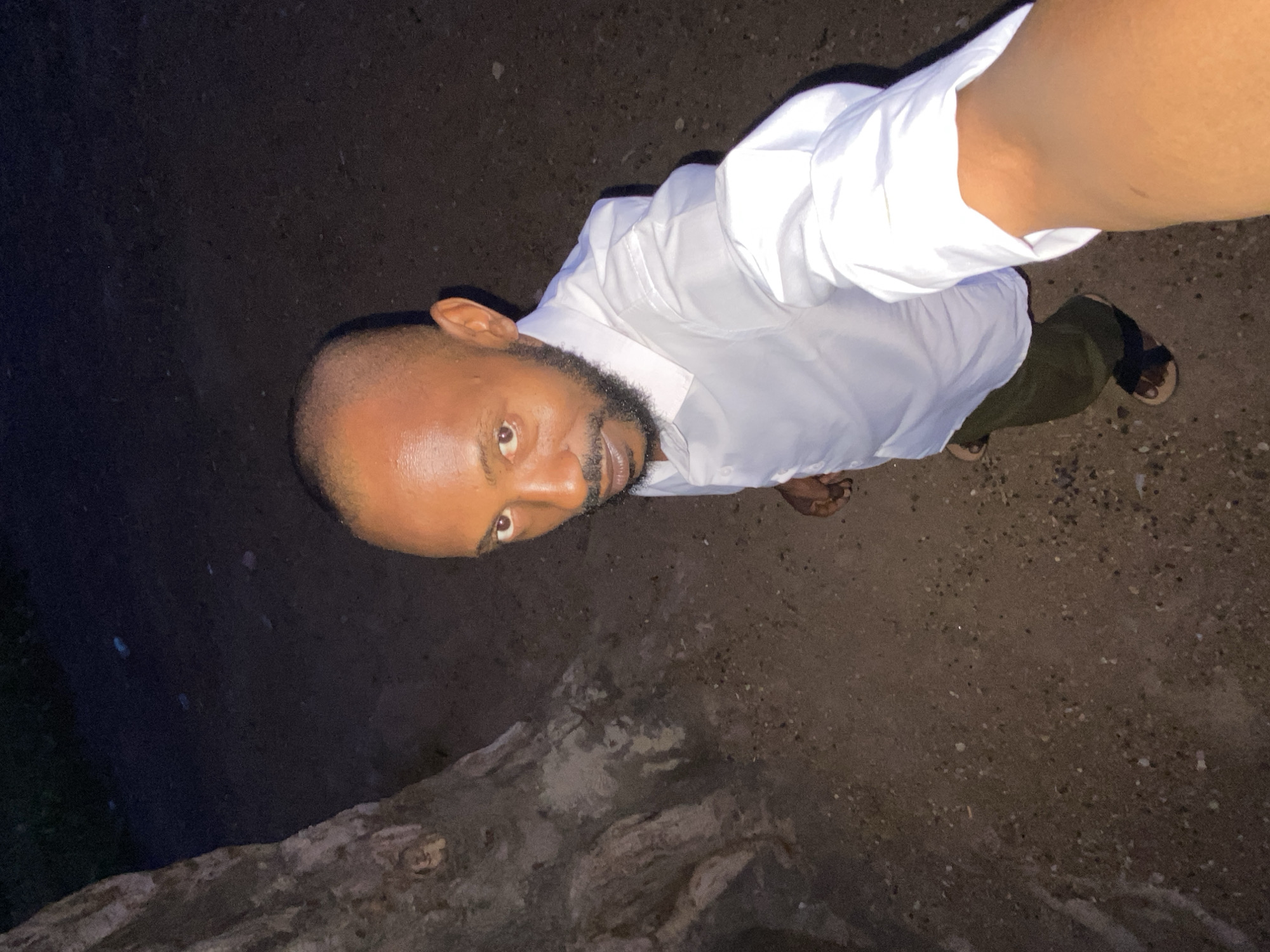 A man is standing outside in Sabon Tasha, Nigeria, on a dirt and gravel surface. He is wearing a white long-sleeved shirt, olive-colored pants, and sandals. He appears to be taking a picture of himself from a low angle. The lighting is dim, with a dark background. A rock or wall is visible in the lower left corner of the frame.