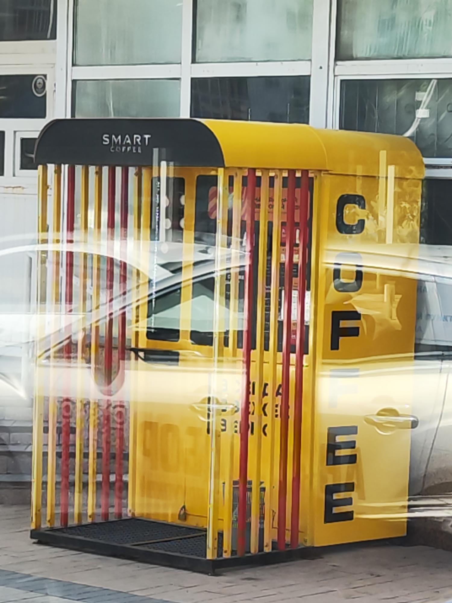 A yellow and black vending machine with vertical red and yellow bars is situated in front of a building with large windows in Astana, Kazakhstan. The machine has the words "SMART COFFEE" written in white on a black panel at the top. On the right side of the machine, large black letters spell out "COFFEE". A white car is visible behind the vending machine, partially obscuring it. The ground in front of the machine is paved with dark grey tiles.