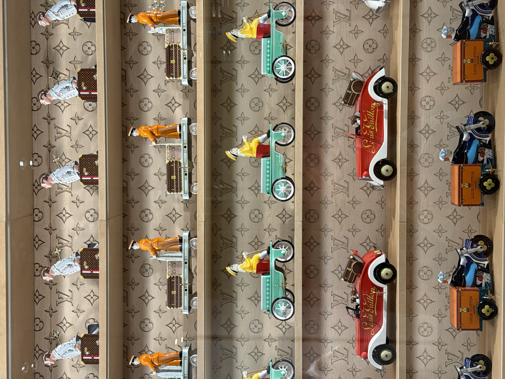 The image shows a display of Louis Vuitton figurines and vehicles, arranged in vertical rows against a background featuring the Louis Vuitton logo. In the first row, there are figurines of men wearing white shirts and carrying luggage. The second row features figurines of men in orange suits standing next to luggage on carts. The third row contains figurines of people driving green, vintage-style cars. The fourth row presents red cars labeled "Louis Vuitton" with trunks carrying luggage. The final row shows figurines with blue shirts and musical instruments on vehicles carrying luggage. The lighting appears to be from above, casting shadows on the display. The display is likely located in Jinjiang District, China.