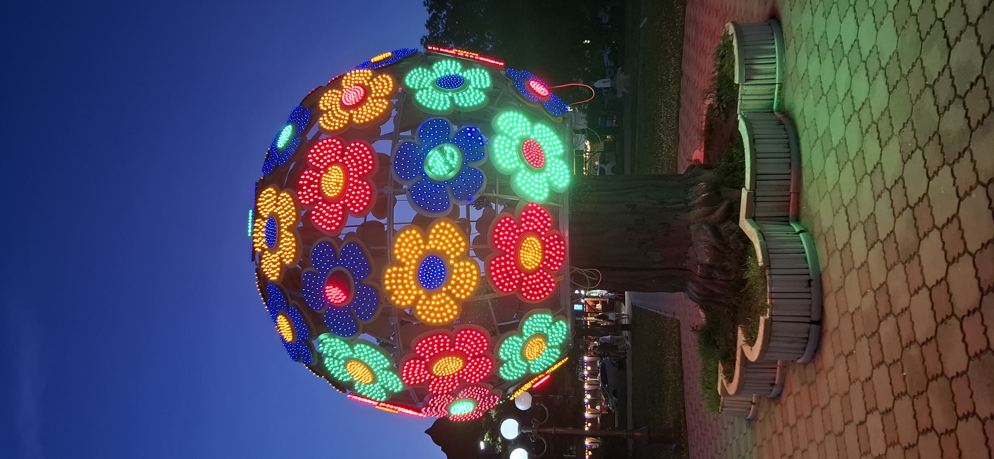 An illuminated, spherical sculpture resembling a giant flower bouquet is centrally located in a park at dusk. The sculpture, made of interconnected flowers, is adorned with numerous LED lights in vibrant colors: red, yellow, blue, and green. Each flower has a central circular area and surrounding petals, all outlined and filled with lights. The sculpture is supported by a large, textured trunk-like structure, which is surrounded by a low, curved planter with grass. The ground is paved with interlocking stone tiles. In the background, a lamppost with multiple lights stands to the left, and beyond that, there is a street with parked cars and a building with illuminated windows. Several people, including adults and children, are visible walking in the background. The sky is a deep blue, indicating twilight. This sculpture is located in Blagoveshchensk, Russia, specifically in the park area near the Amur River embankment.