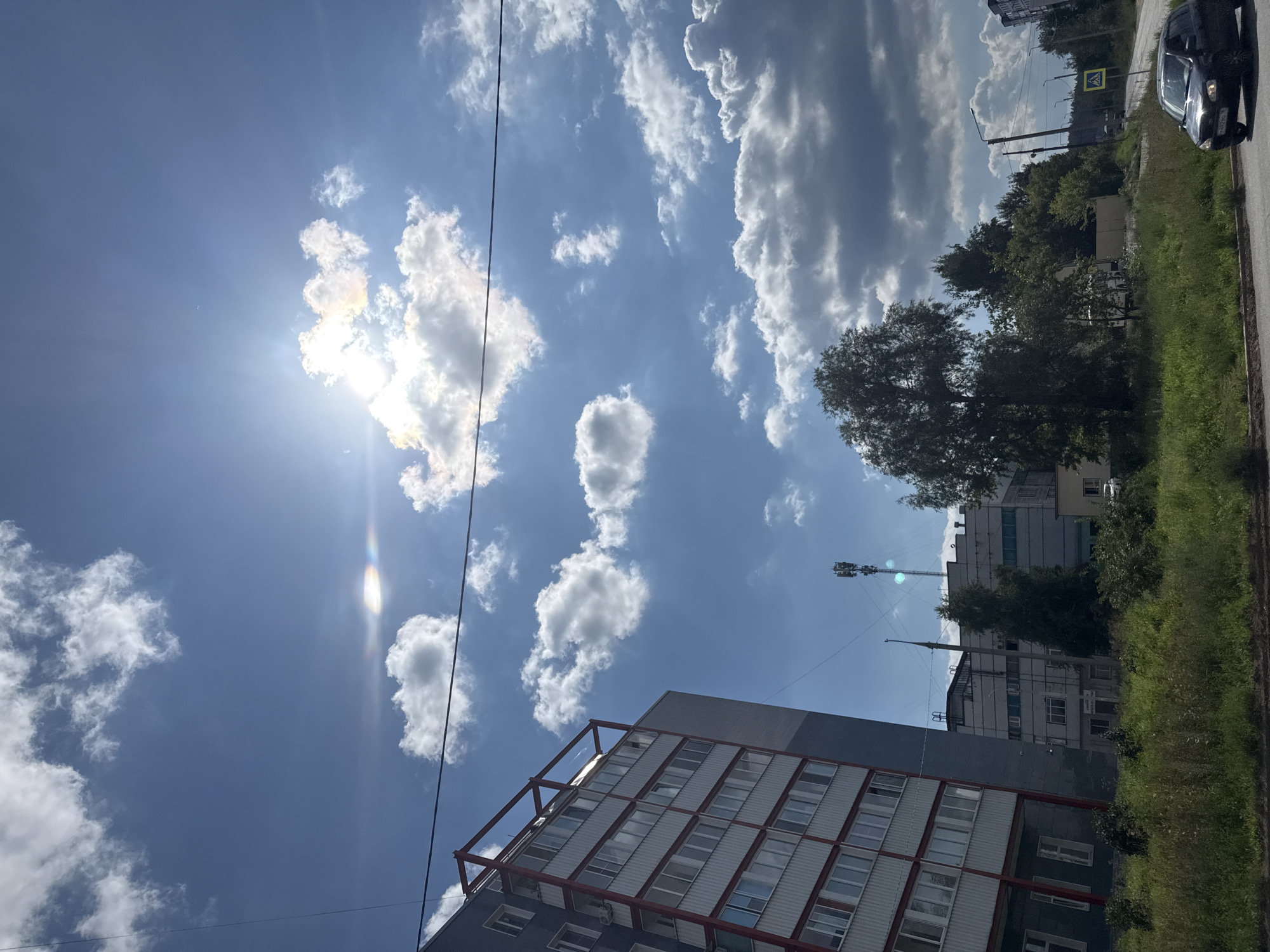 This image depicts a street scene in Chaikovsky, Russia, on a bright, sunny day with scattered clouds. In the foreground, a dark-colored car is driving on a paved road. To the left, a multi-story apartment building with a grey facade and white balconies trimmed in red is visible. Behind the apartment building, there are other buildings and a telecommunications tower. A line of trees and overgrown green grass separates the road from the buildings. The sky is a vivid blue with fluffy white clouds, and the sun is shining brightly, creating a slight lens flare and rainbow effect. A yellow pedestrian crossing sign is present on the right side of the road.