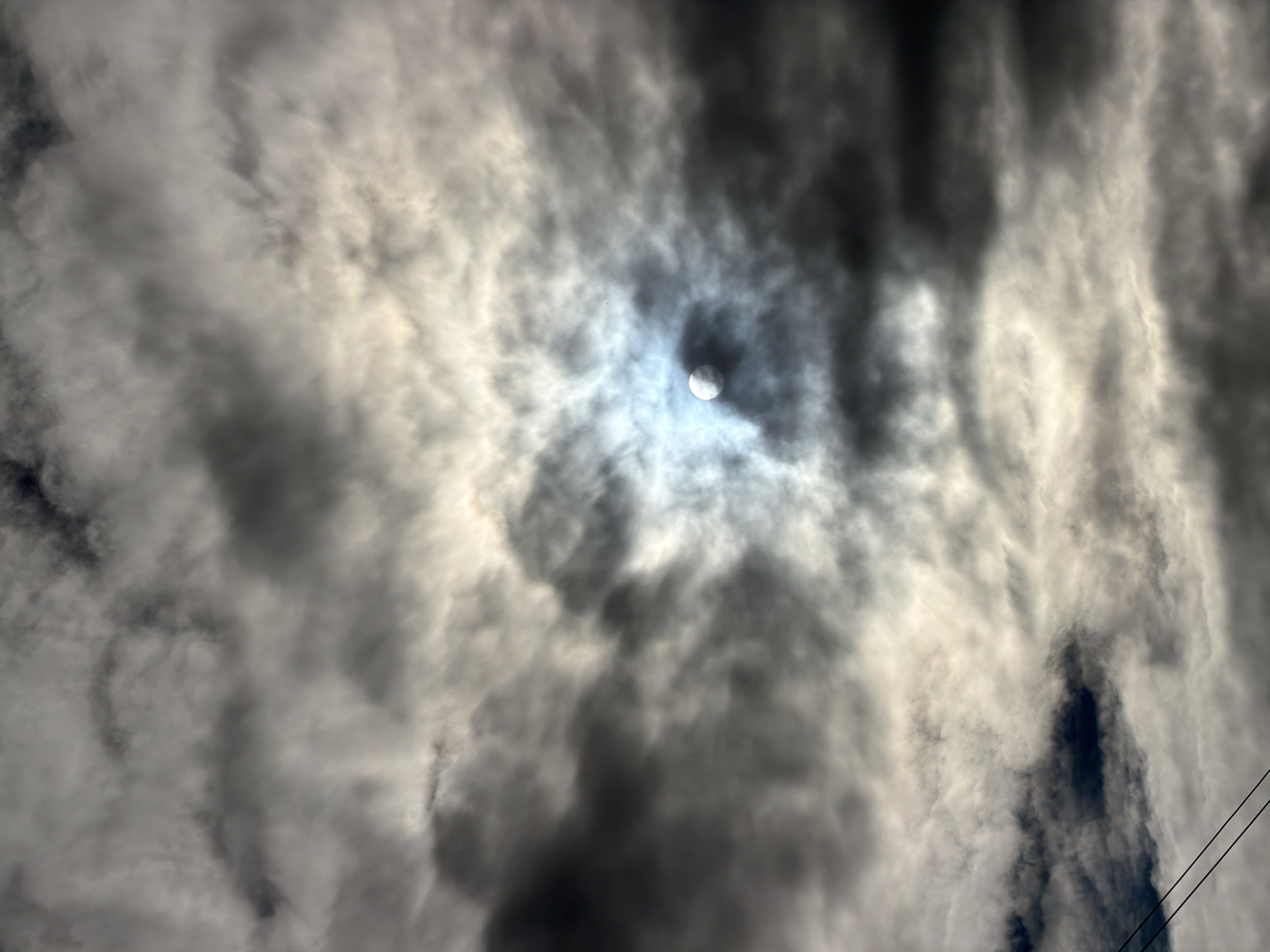 The media file shows a dramatic sky filled with dark, swirling clouds. The sun is partially visible through a gap in the clouds, appearing as a bright, ethereal disc surrounded by a halo of light. The overall impression is one of a dynamic and powerful sky, possibly indicating an approaching storm or the aftermath of one. The light filtering through the clouds creates a contrast between light and shadow, adding to the dramatic effect. In the lower right corner, two thin, dark lines that appear to be power lines are visible, suggesting an outdoor scene, possibly in a rural or suburban setting. There is no indication of people or specific objects in the scene, other than the power lines. The time of day is not explicitly stated, but the lighting suggests it could be late afternoon or early evening, when the sun is lower in the sky. The weather appears to be overcast with breaks of sunlight. The location context of Moshenskoe, Russia, does not provide specific visual cues within the image itself, but suggests a natural, potentially vast landscape. The emotions evoked by the image are a sense of awe, power, and perhaps a touch of foreboding due to the dark clouds. There is no visible text in the media file.