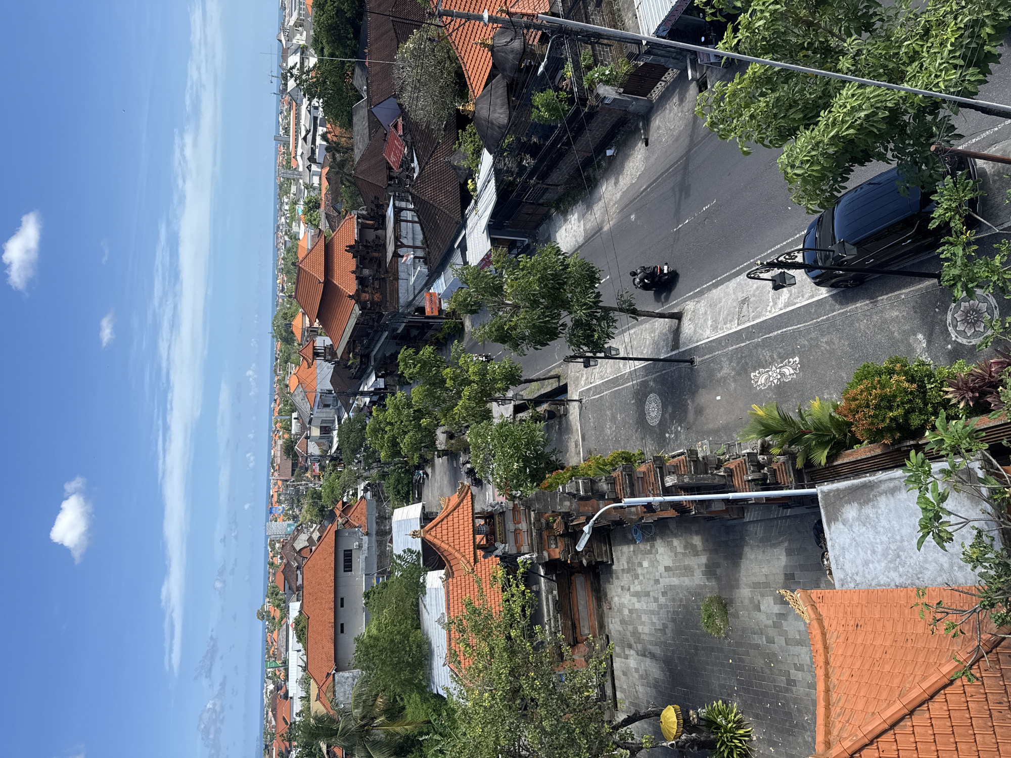 The image shows an aerial view of a street in North Kuta, Indonesia. The street is lined with buildings with red tiled roofs and lush green trees. A black car is parked on the right side of the road, and a motorcycle is driving down the street. The sky is blue with some clouds.