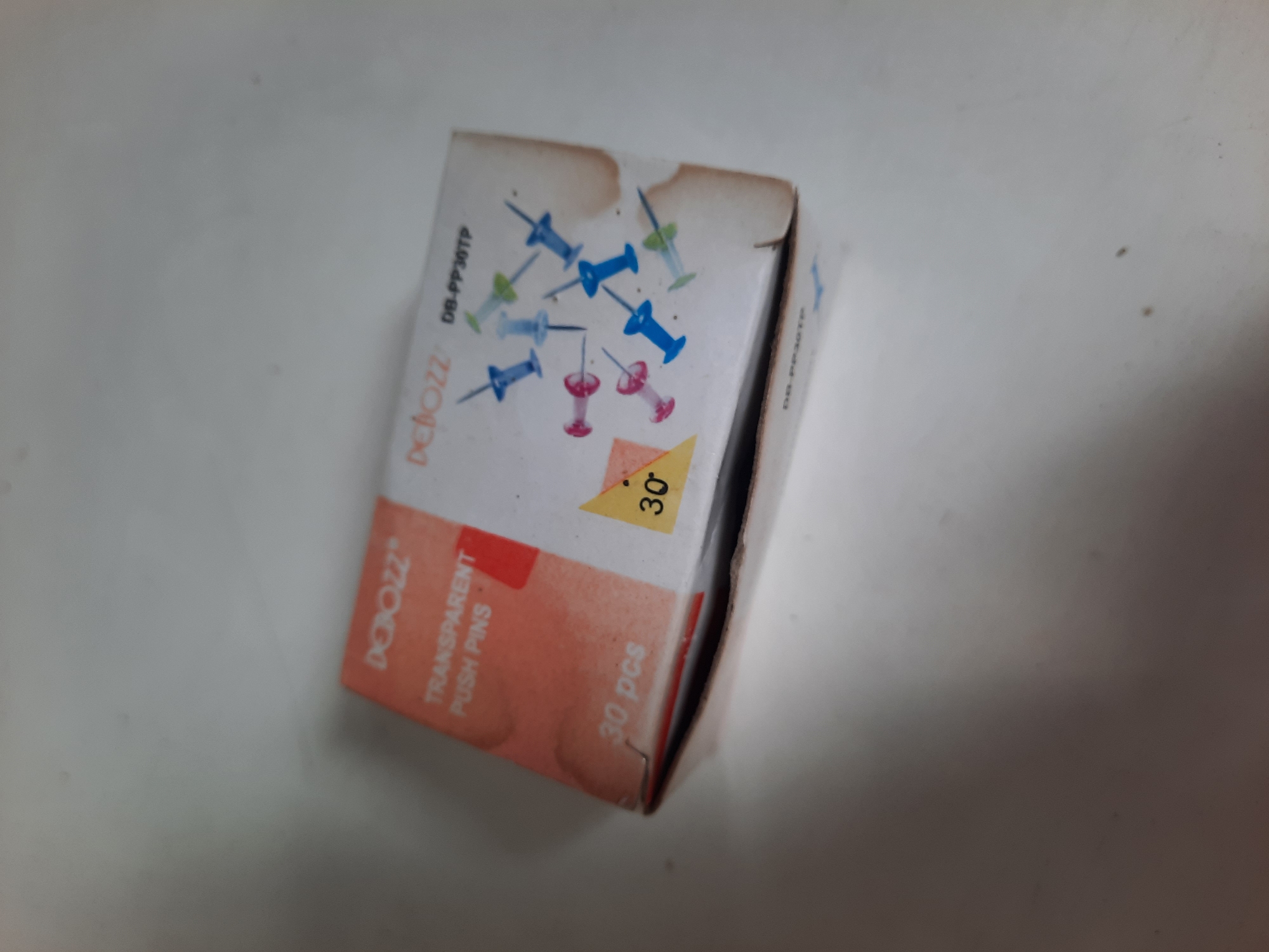 A partially opened box of transparent push pins is on a white surface. The box is orange and white, with the brand name "DEBOZZ" and "TRANSPARENT PUSH PINS" printed on it. There are illustrations of colorful push pins on the side of the box. The box also states "30 pcs". The surface the box is on appears to be a table or countertop. The image is taken from a slightly elevated angle. There is no visible context of Lhokseumawe, Indonesia in the image.
